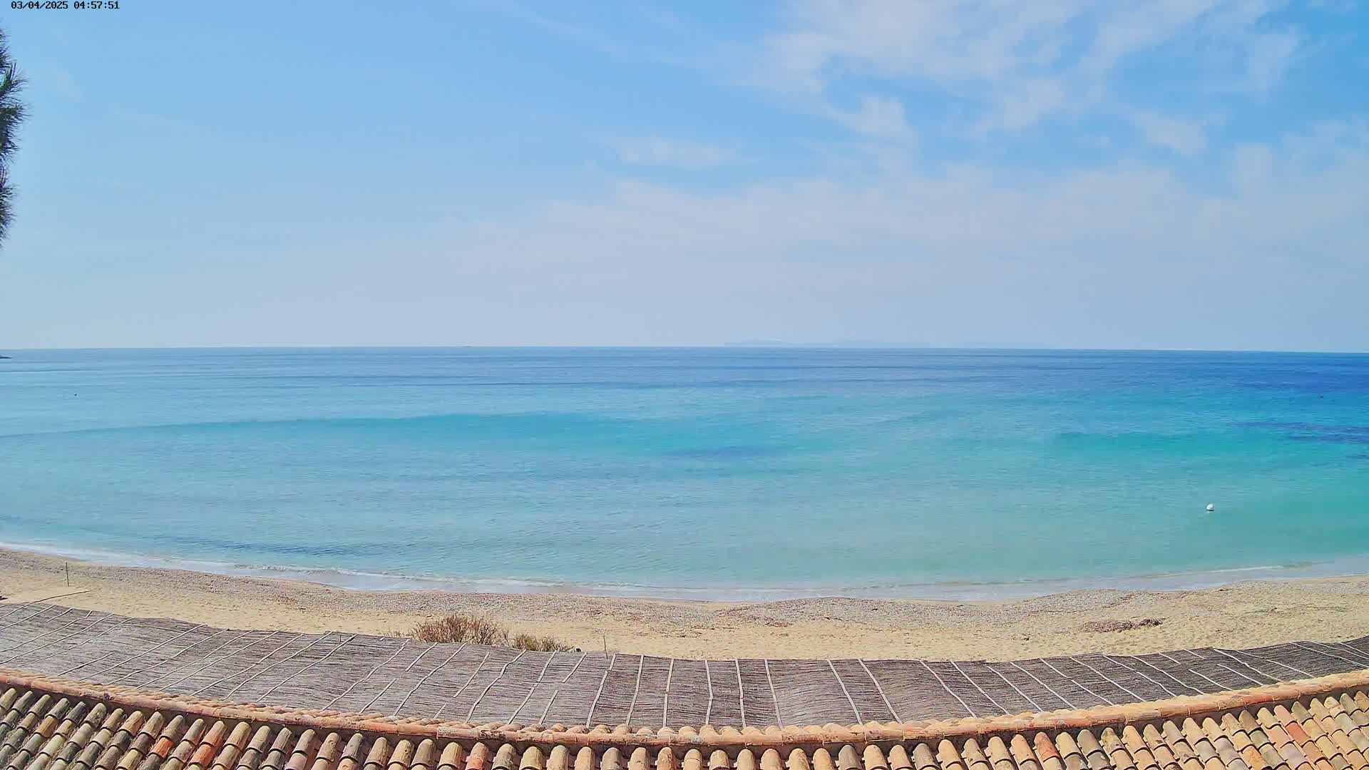 A sandy beach with calm, turquoise water under a mostly clear, sunny sky is viewed from above a tiled roof.