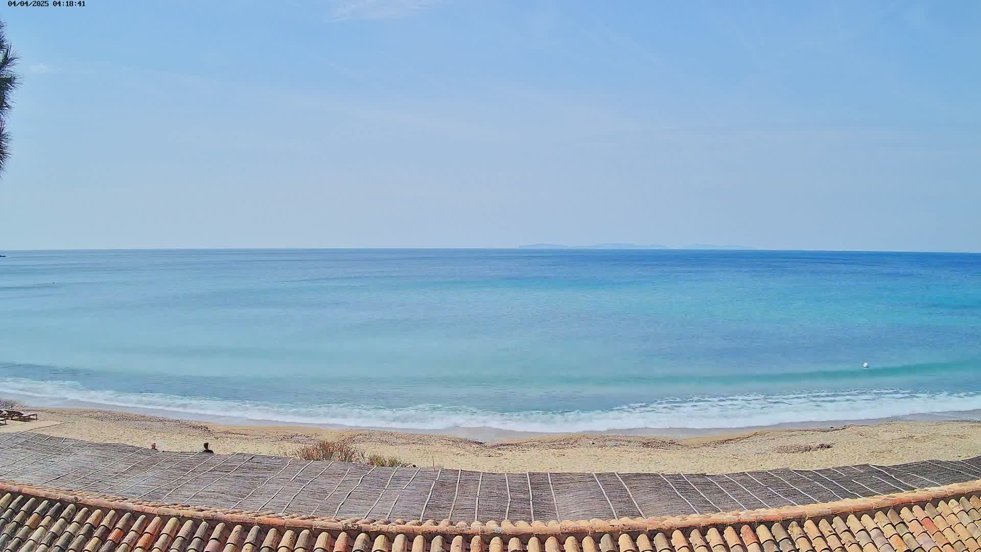 A sandy beach with gentle waves laps the shore of a calm, light-blue ocean under a clear blue sky.