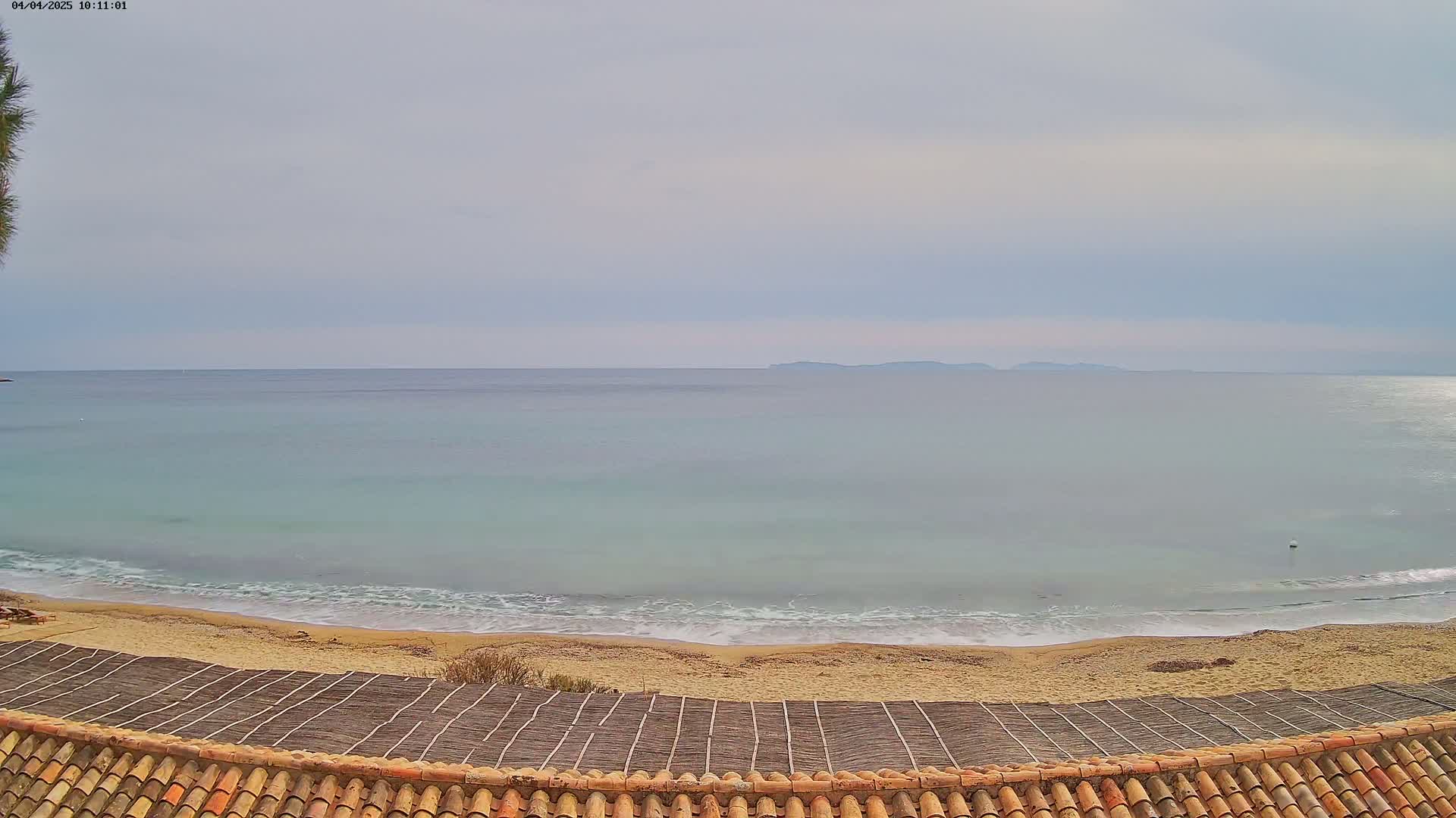 A sandy beach with gentle waves meets a calm, light-blue sea under a cloudy sky, and a distant island is visible on the horizon.