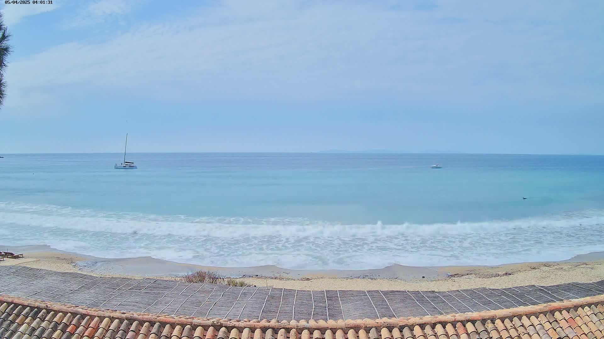 A tranquil beach scene under a mostly clear sky shows a calm sea with small waves lapping a sandy shore, and a few boats visible in the distance.