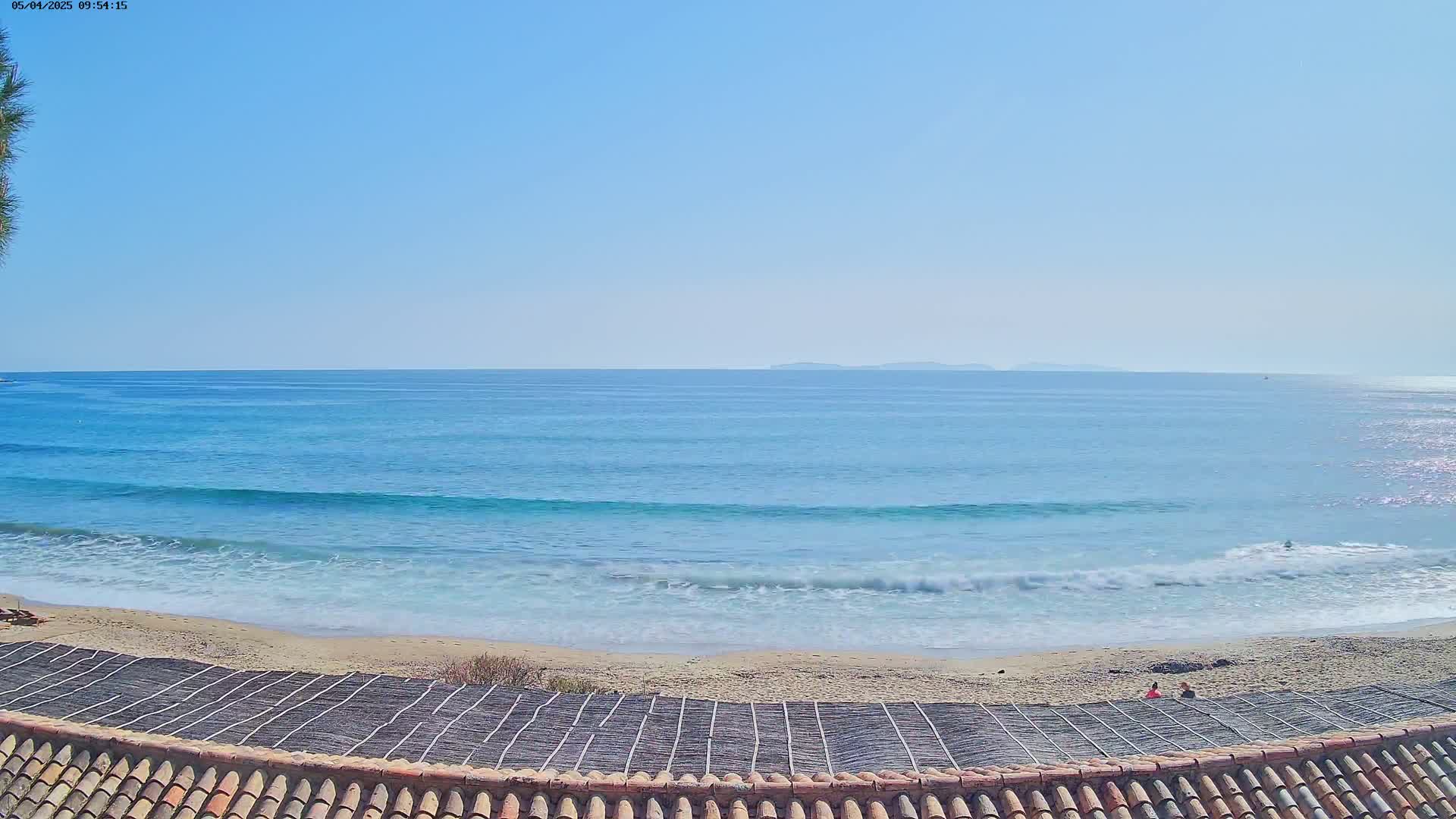 A sandy beach with small waves under a clear blue sky is seen from above a tiled roof.