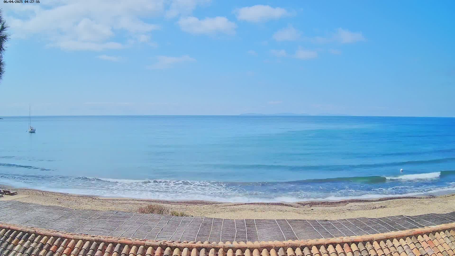 A sandy beach with gentle waves laps the shore under a mostly sunny sky, with a sailboat visible in the distance.