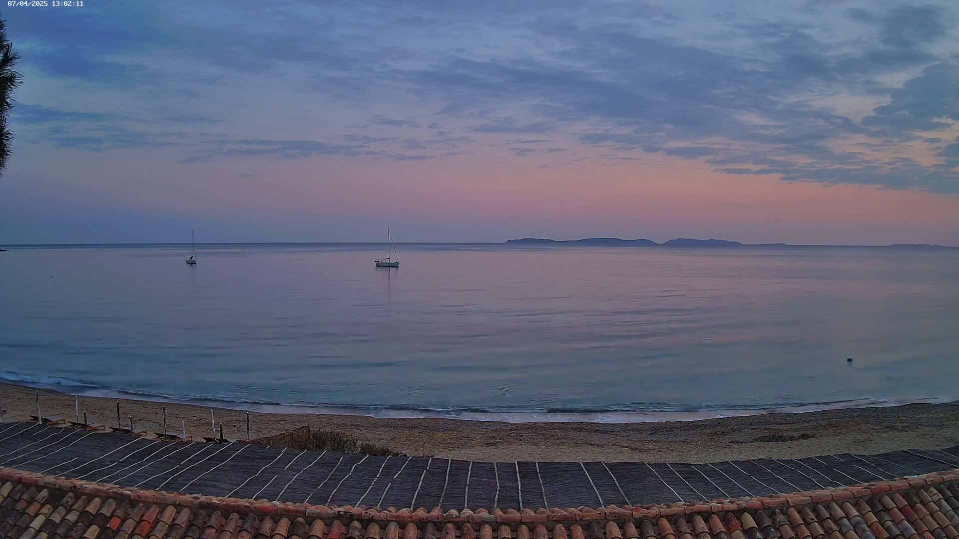 A calm, pale pink and blue sunset over a tranquil sea, with a few sailboats and a sandy beach visible from a tiled roof.