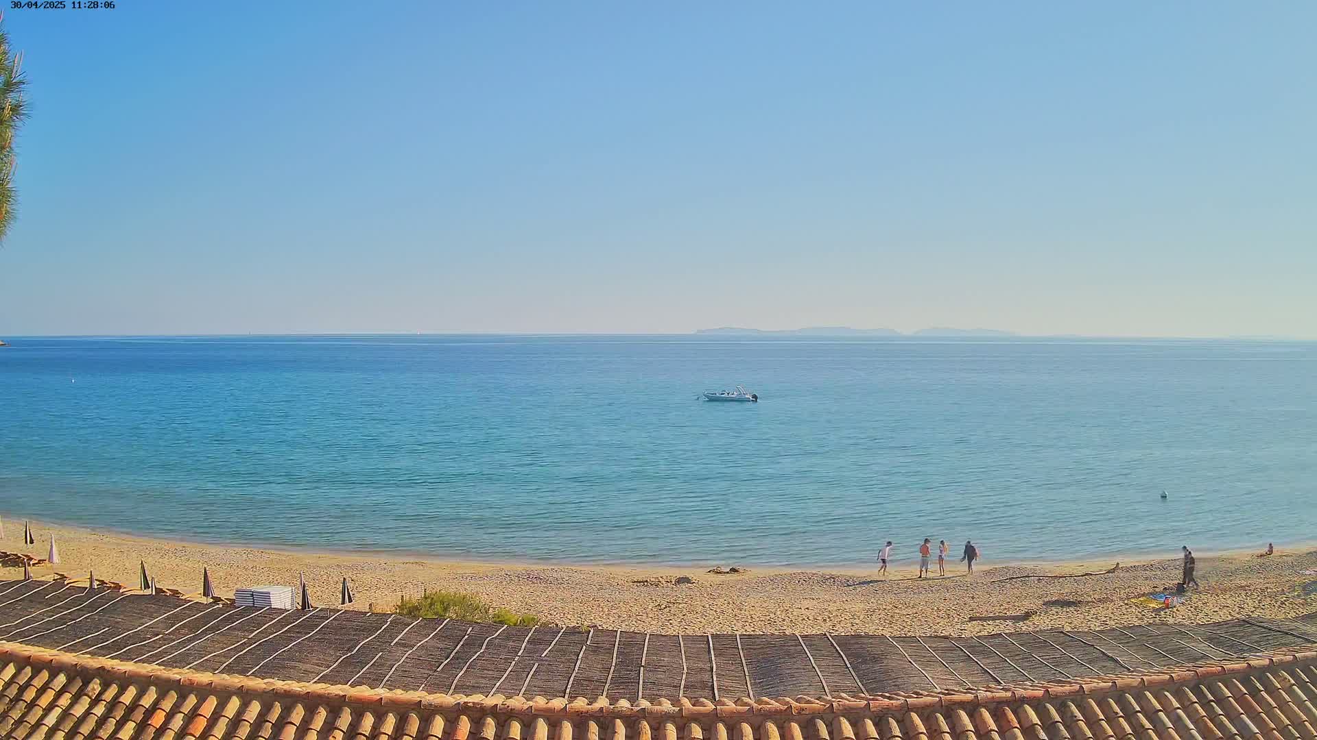 A sandy beach with a few people on it slopes down to a calm, clear blue sea under a bright, sunny sky, with a small boat visible on the water and land visible in the distance.