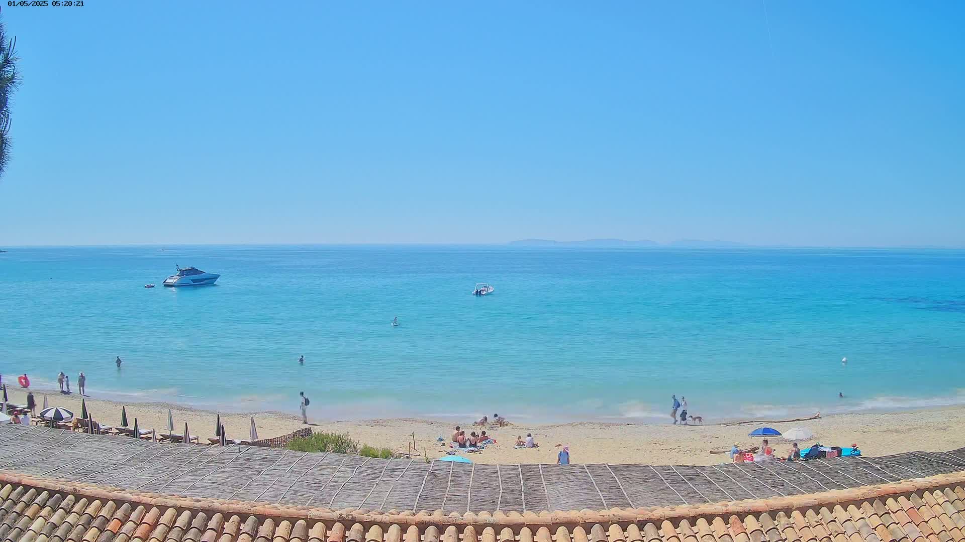A sandy beach with several people relaxing under umbrellas and in the water, a boat offshore, and a clear blue sky on a sunny day.