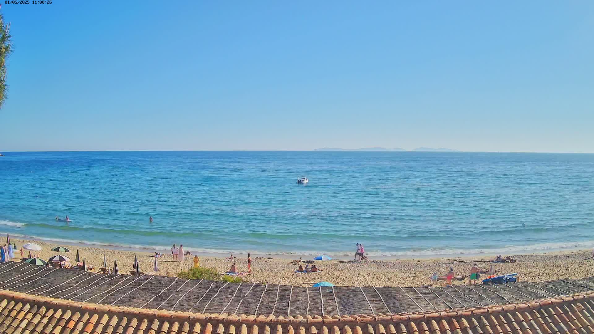 A sandy beach with scattered beachgoers and umbrellas under a clear blue sky overlooks a calm, turquoise ocean with a small boat visible in the distance.