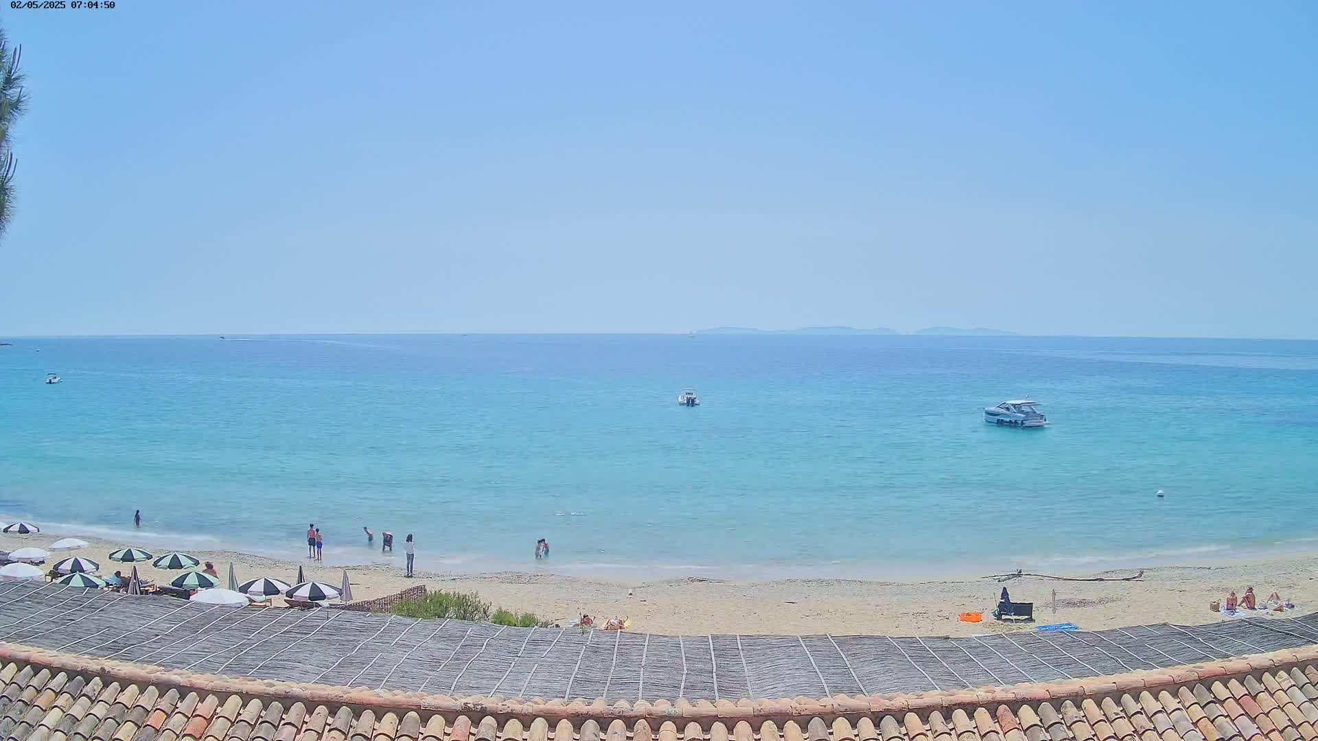 A sandy beach with a few people and umbrellas is visible under a clear blue sky, with calm turquoise water and several small boats in the distance.