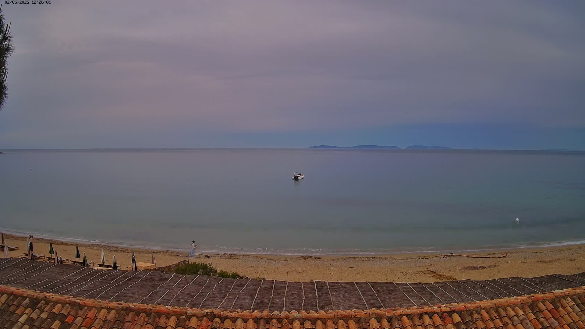A calm, overcast day shows a sandy beach with a few people, a small boat on the water, and distant land visible across a quiet sea.