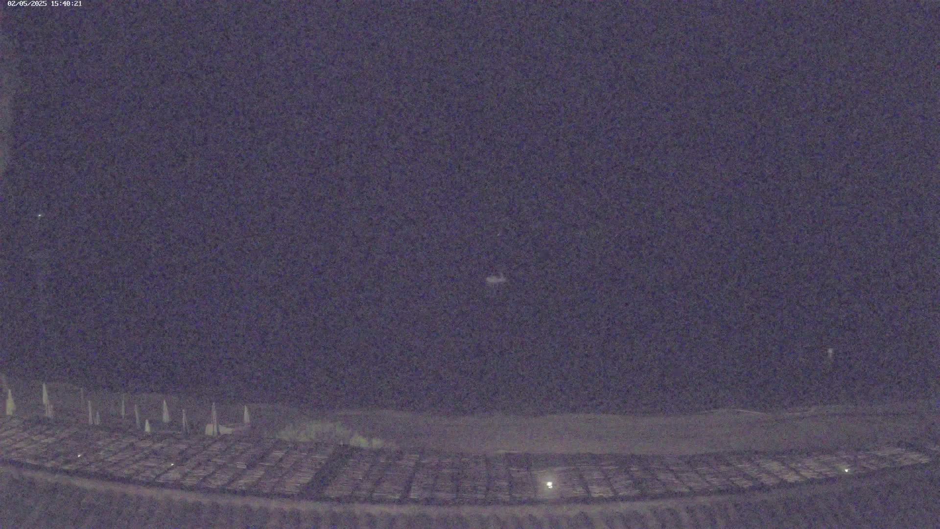 A dark, nighttime scene shows a large, flat area with rows of objects, possibly parked vehicles or equipment, under a clear, dark sky.