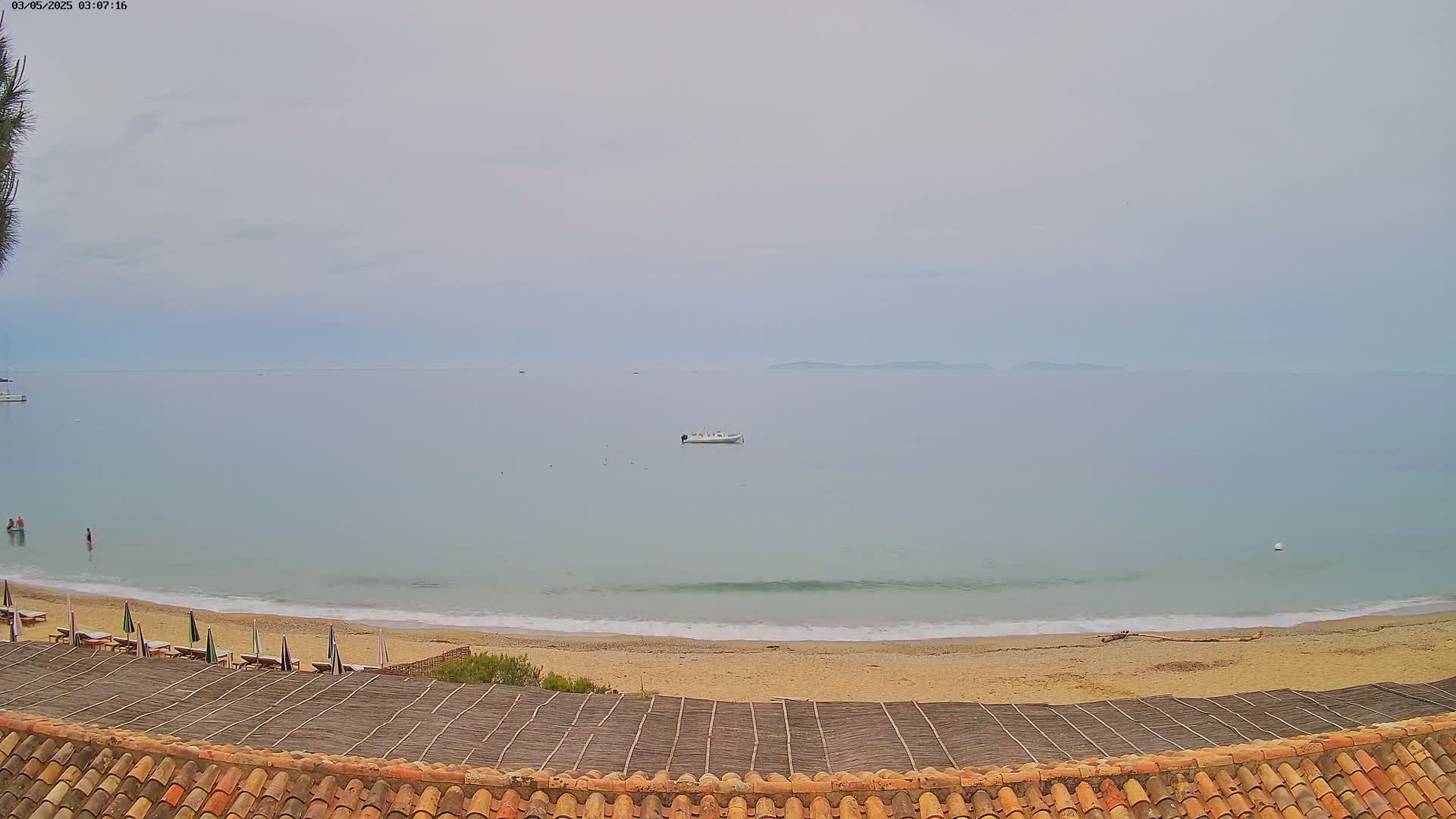 A sandy beach with calm, light-blue water and a few people swimming, is viewed from above an orange-tiled roof under an overcast sky.