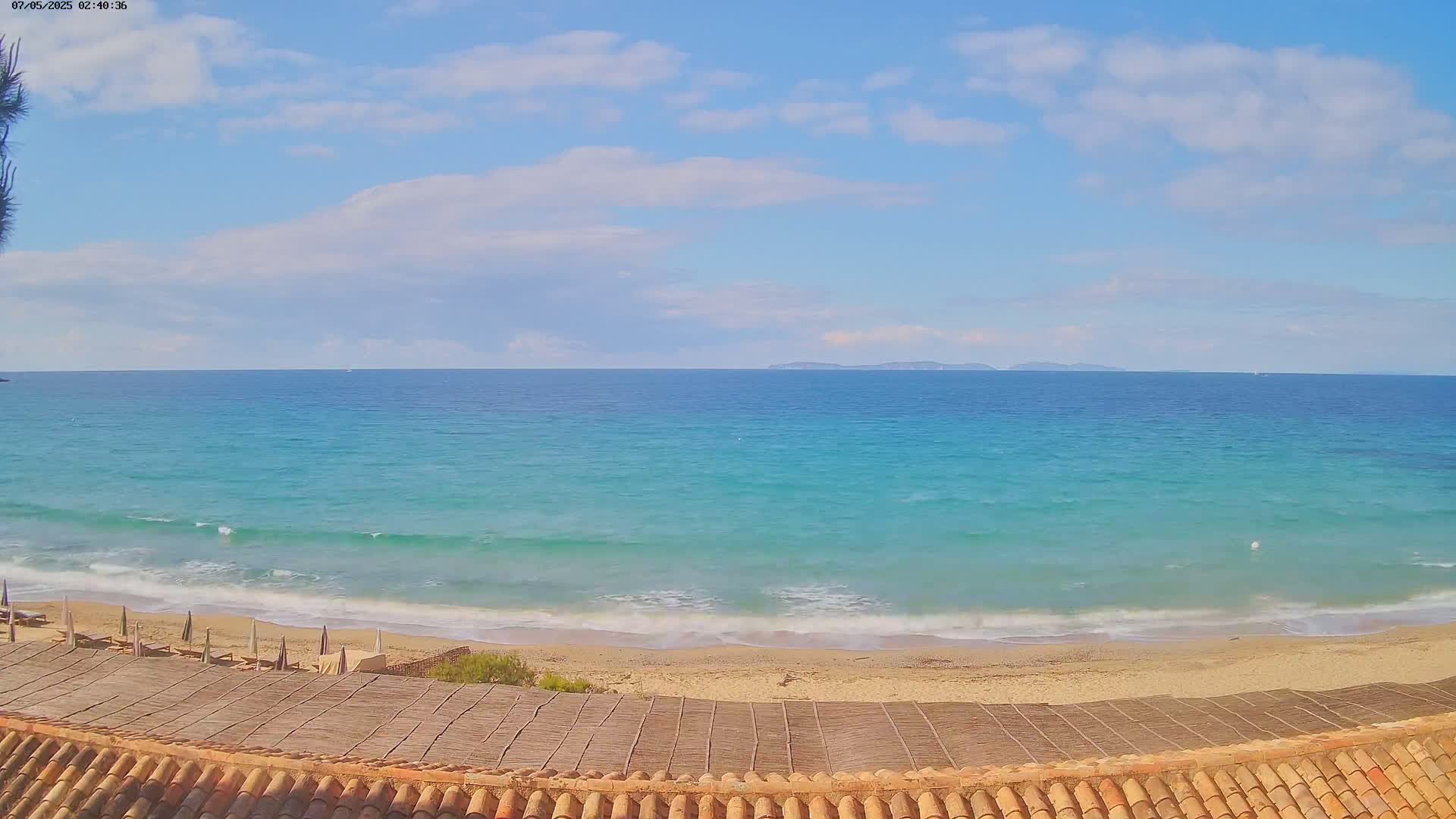 A sandy beach with a turquoise sea and a partly cloudy sky is seen from a vantage point above a thatched roof.