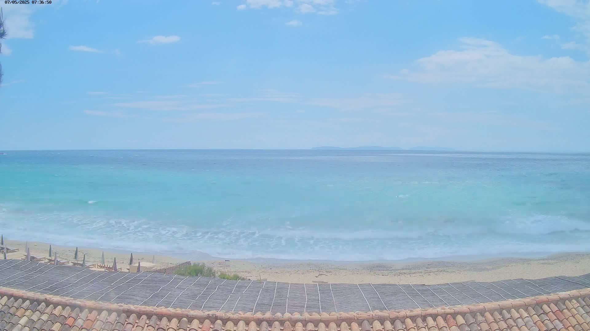 A sandy beach with light waves and a turquoise sea under a mostly clear sky is viewed from a roof with a thatched covering.