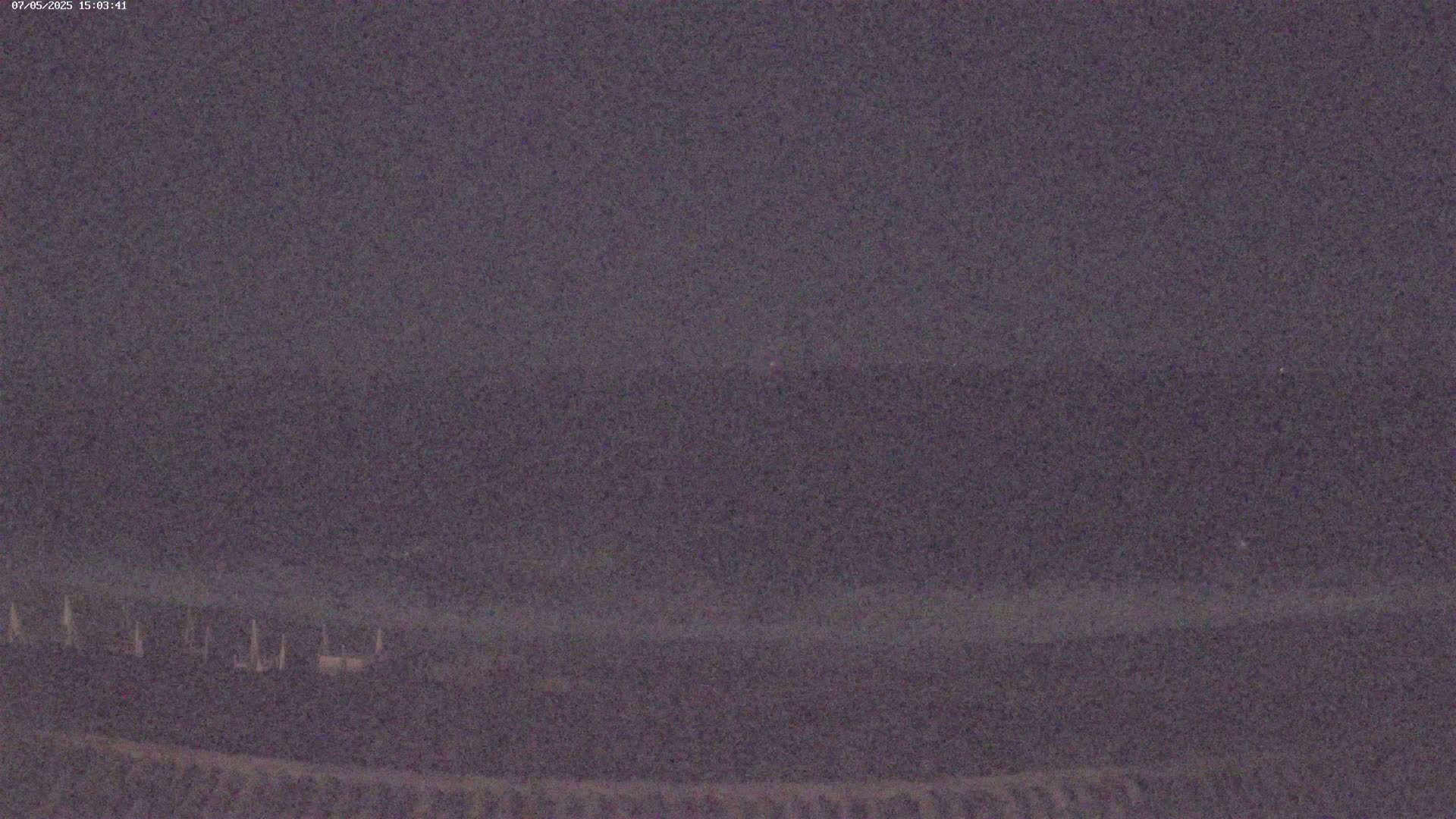 A dark, blurry nighttime image shows a low horizon line with indistinct shapes possibly representing objects or structures in the distance under a dark sky.