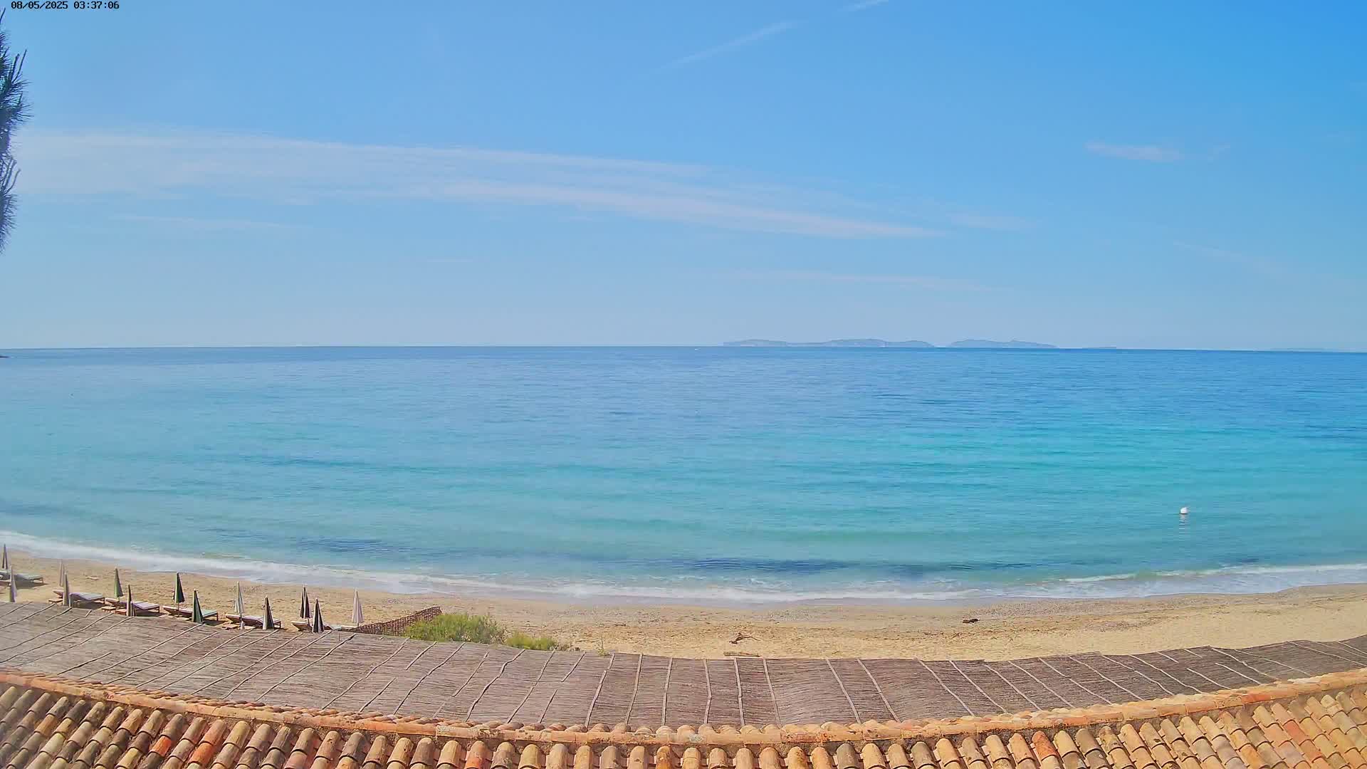 A sandy beach with calm, turquoise water and a few closed umbrellas under a mostly clear blue sky stretches to distant islands.