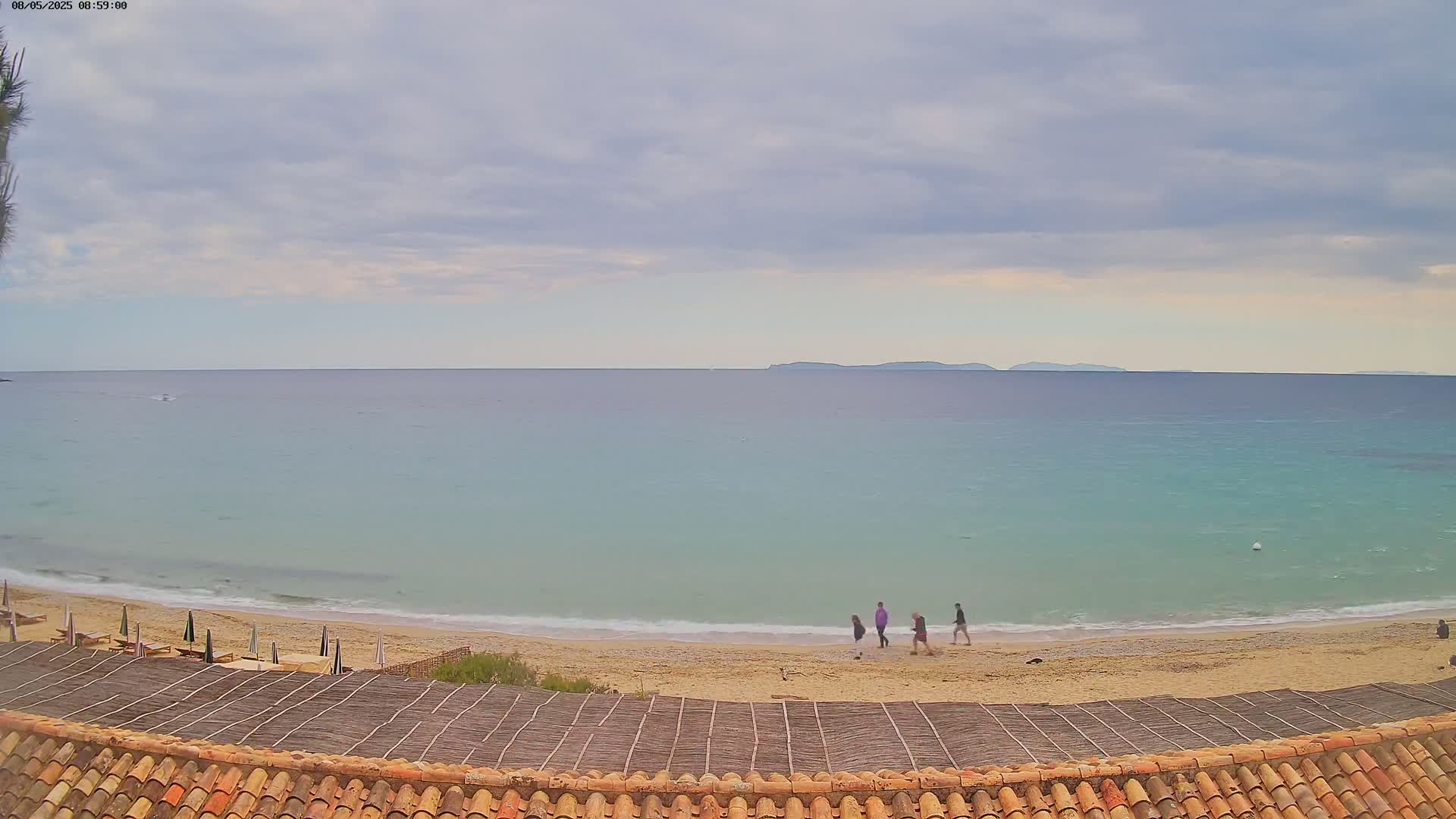 A sandy beach with a few people walking along the shore and calm, light-blue water under an overcast sky, with distant islands visible on the horizon.