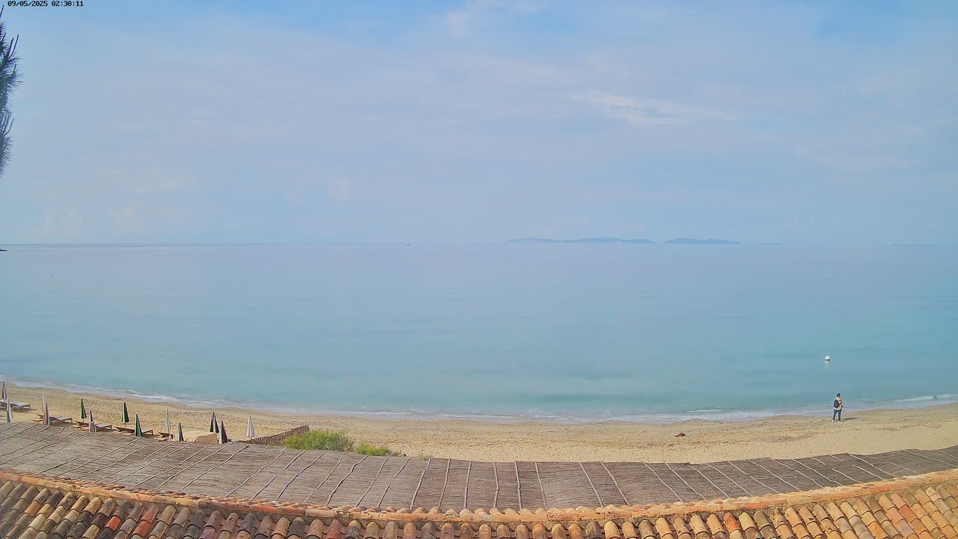A sandy beach with a few closed umbrellas and a person walking on it slopes down to a calm, light-blue ocean under a mostly clear sky.