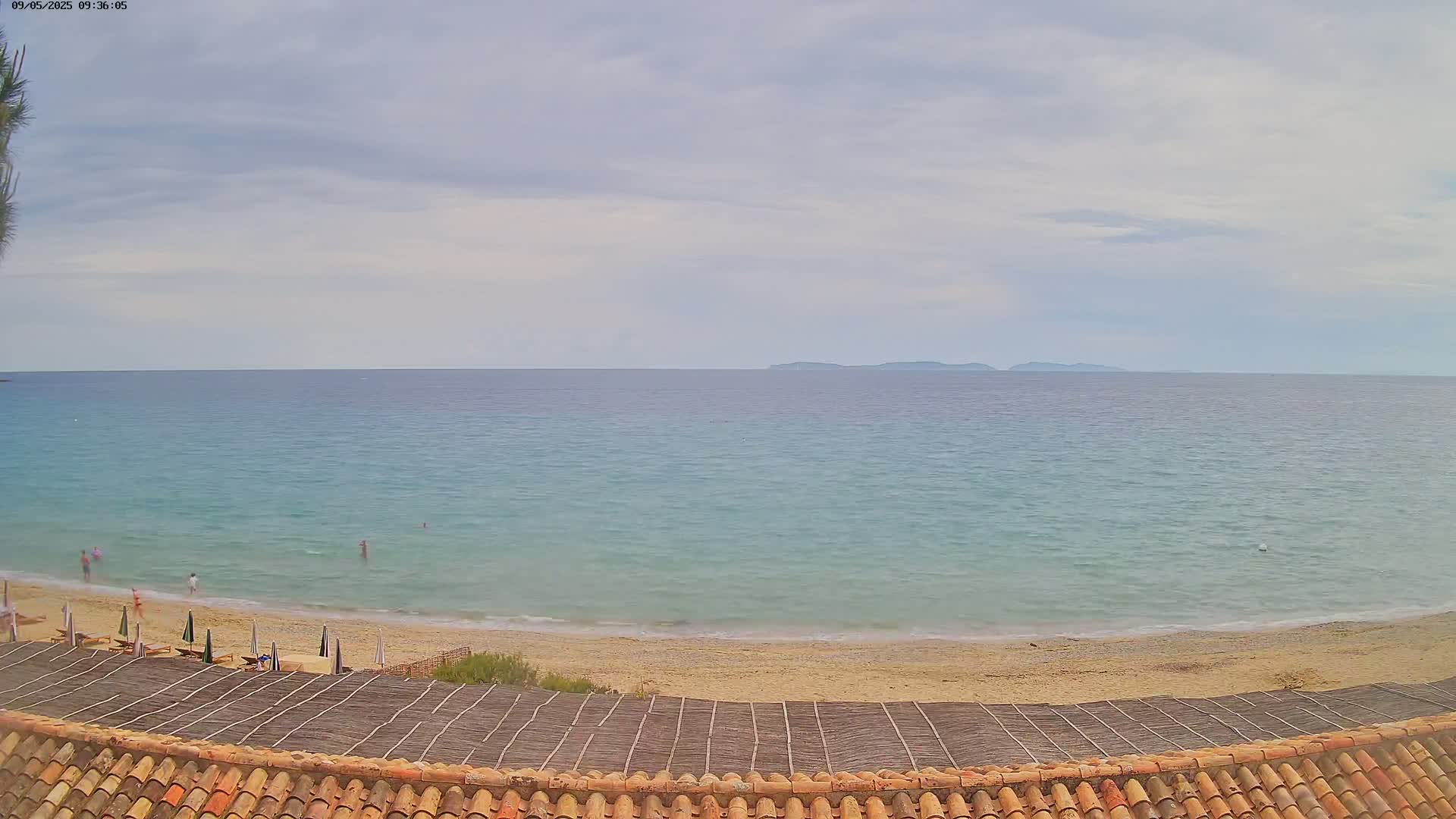 A mostly sunny beach scene shows a calm, light-blue sea with a distant island under a cloudy sky, viewed from a tiled roof above a sandy beach with a few people and beach umbrellas.
