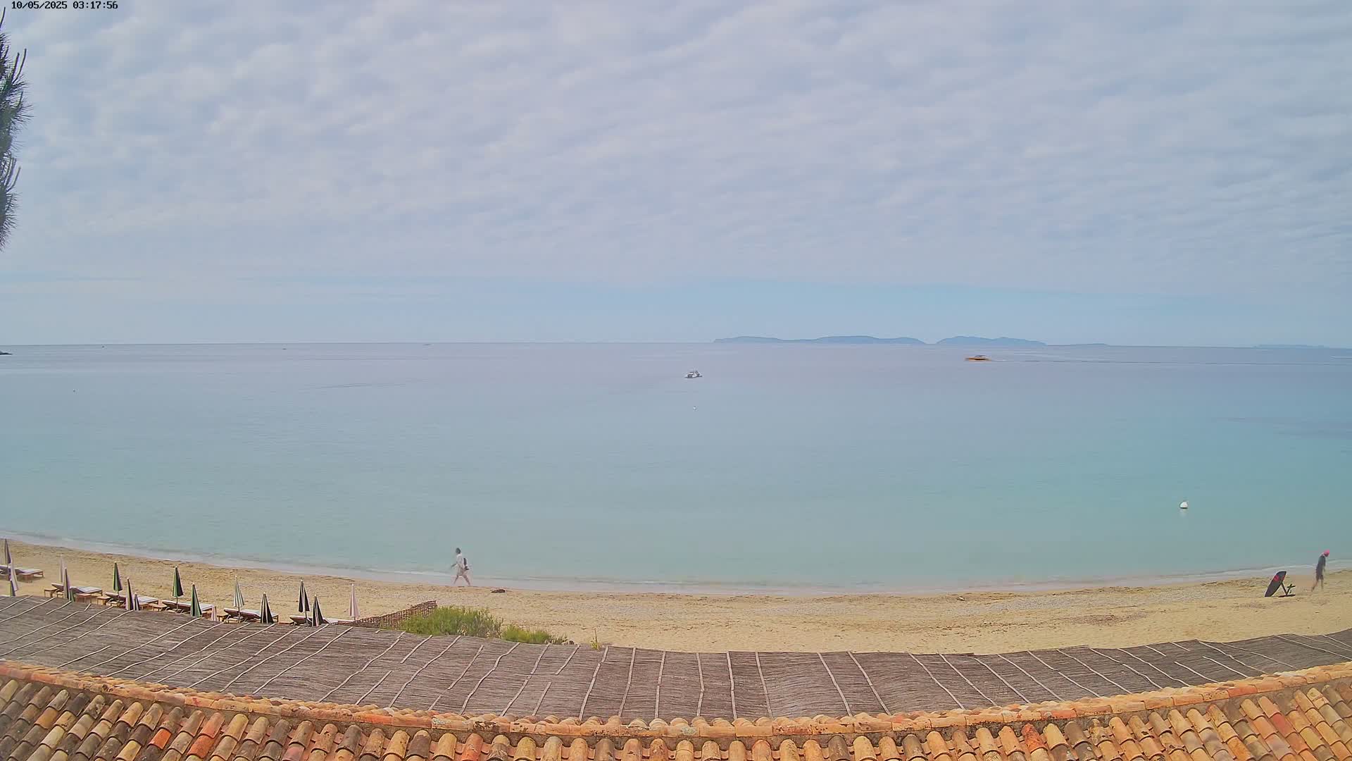 A sandy beach with a few people and lounge chairs stretches along a calm, light-blue sea under a mostly cloudy sky.