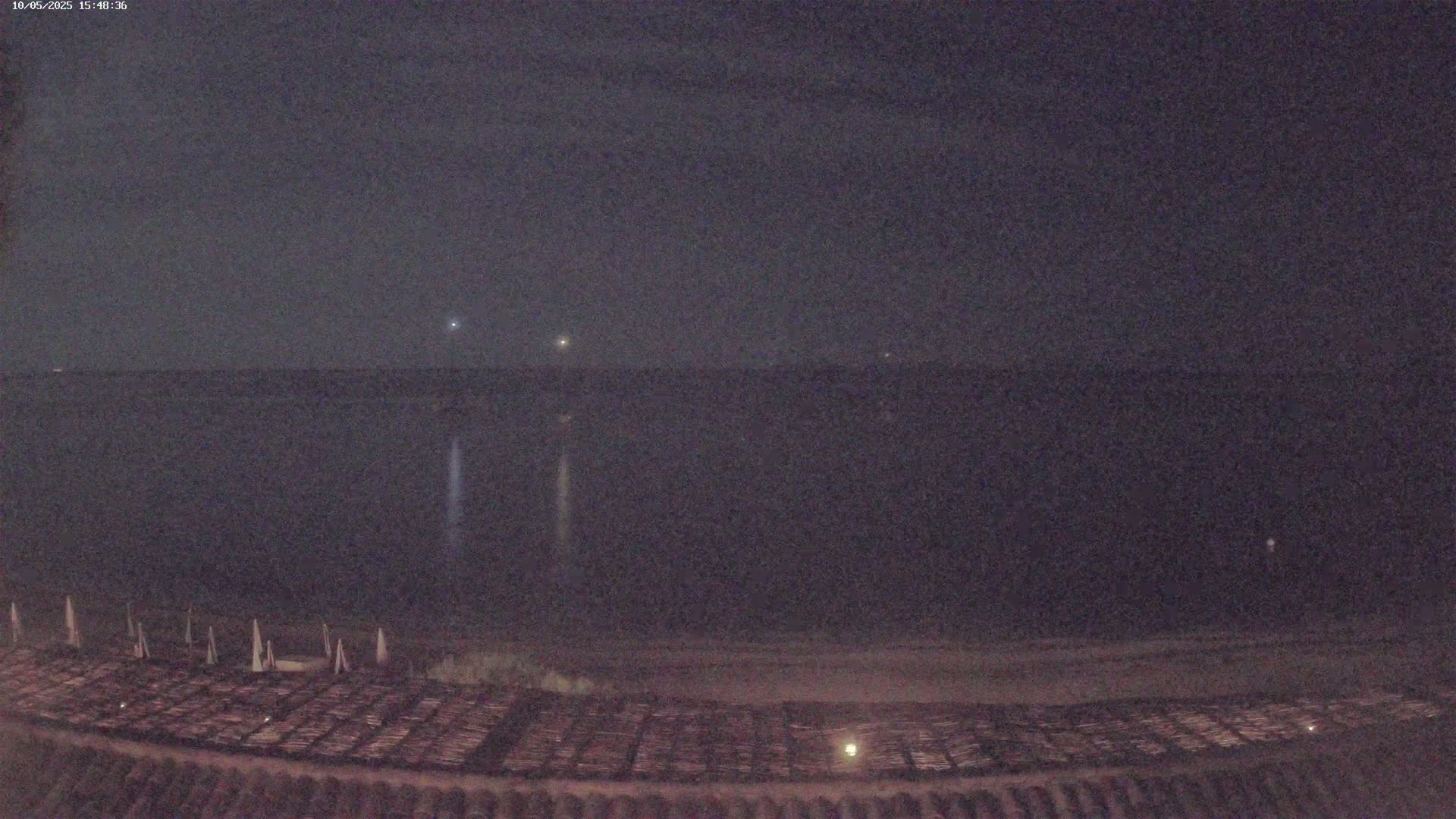 A dark, nighttime beach scene shows a calm body of water under a mostly clear sky, with a few faint lights visible in the distance and folded beach furniture in the foreground.