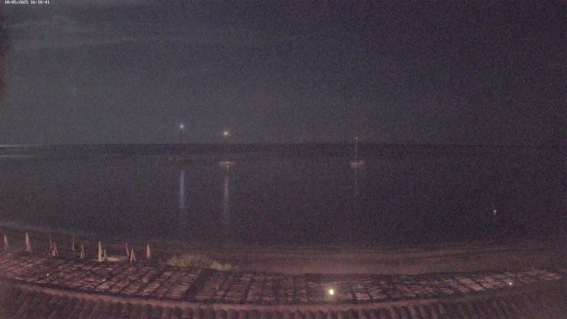 At night, a dark body of water with a few boats is visible from a beach with rows of what appear to be beach umbrellas laid out on the sand.