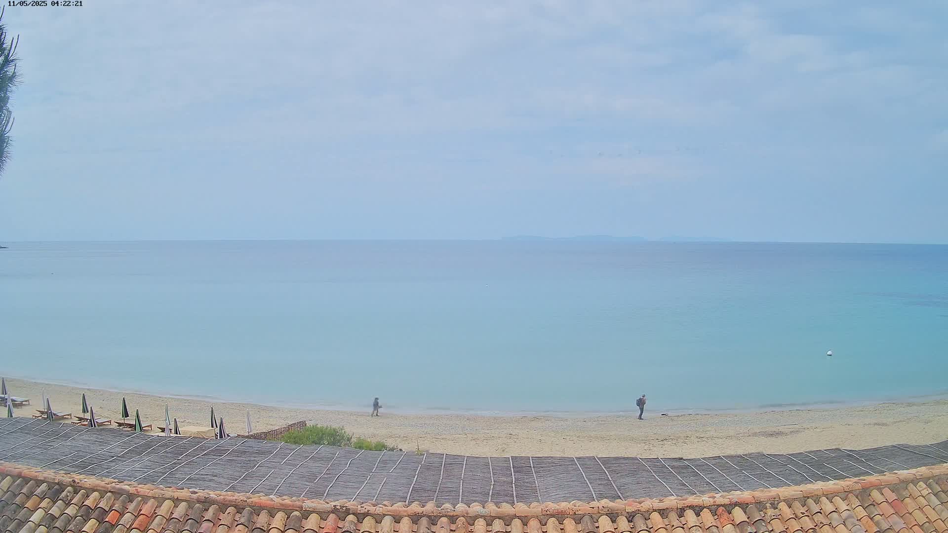 A sandy beach with a few people and several closed umbrellas stretches along a calm, light-blue sea under an overcast sky.