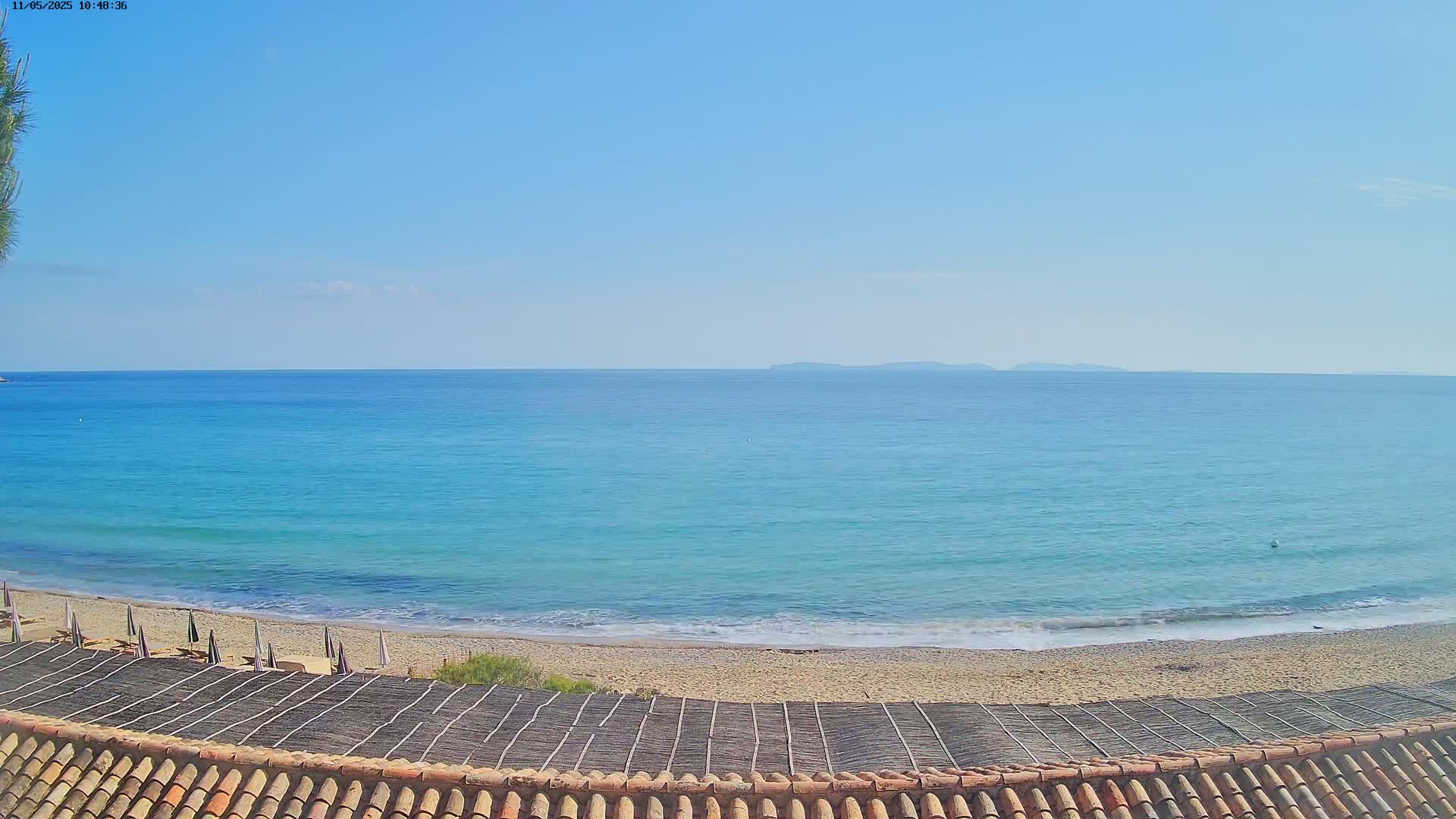 A sandy beach with calm, turquoise water and a distant landmass under a clear blue sky is viewed from a tiled roof.