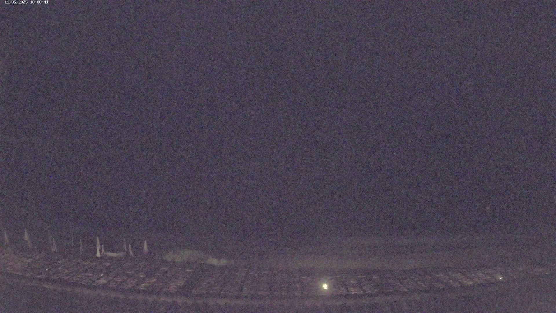 A nighttime, low-light view shows a dark, flat expanse with indistinct, light-colored objects in the distance, possibly structures or markers, under a dark, overcast sky.