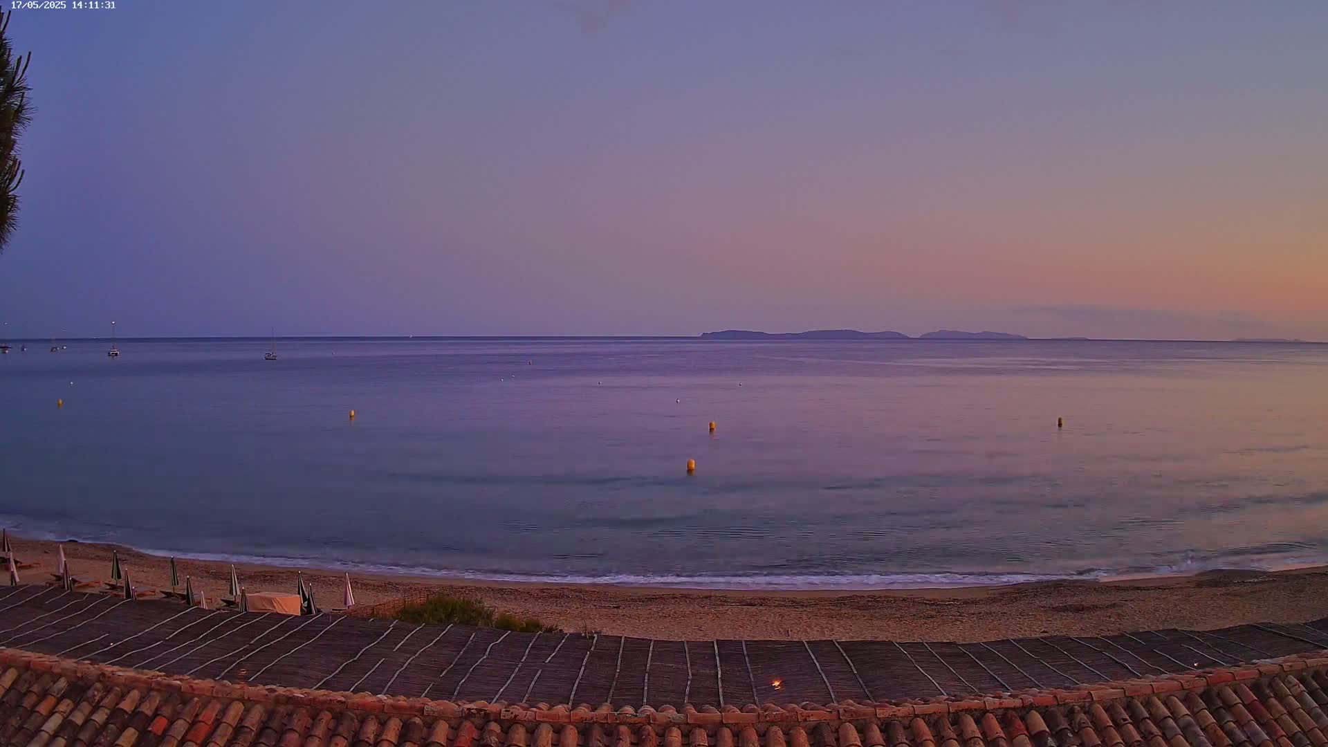 A tranquil beach scene at dusk shows a calm sea under a purple and pink sky, with a few boats and buoys visible in the distance.