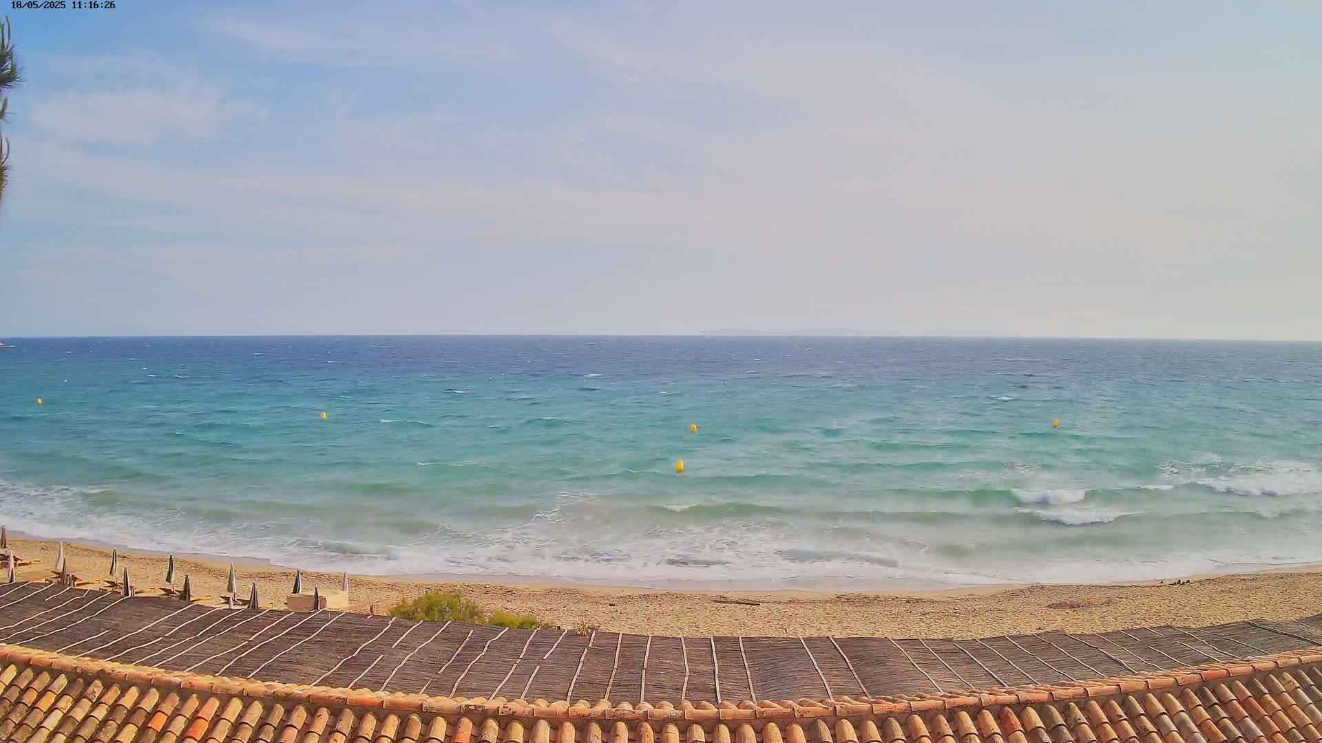 A mostly sunny day shows a wavy teal ocean meeting a sandy beach, viewed from a roof with terracotta tiles.