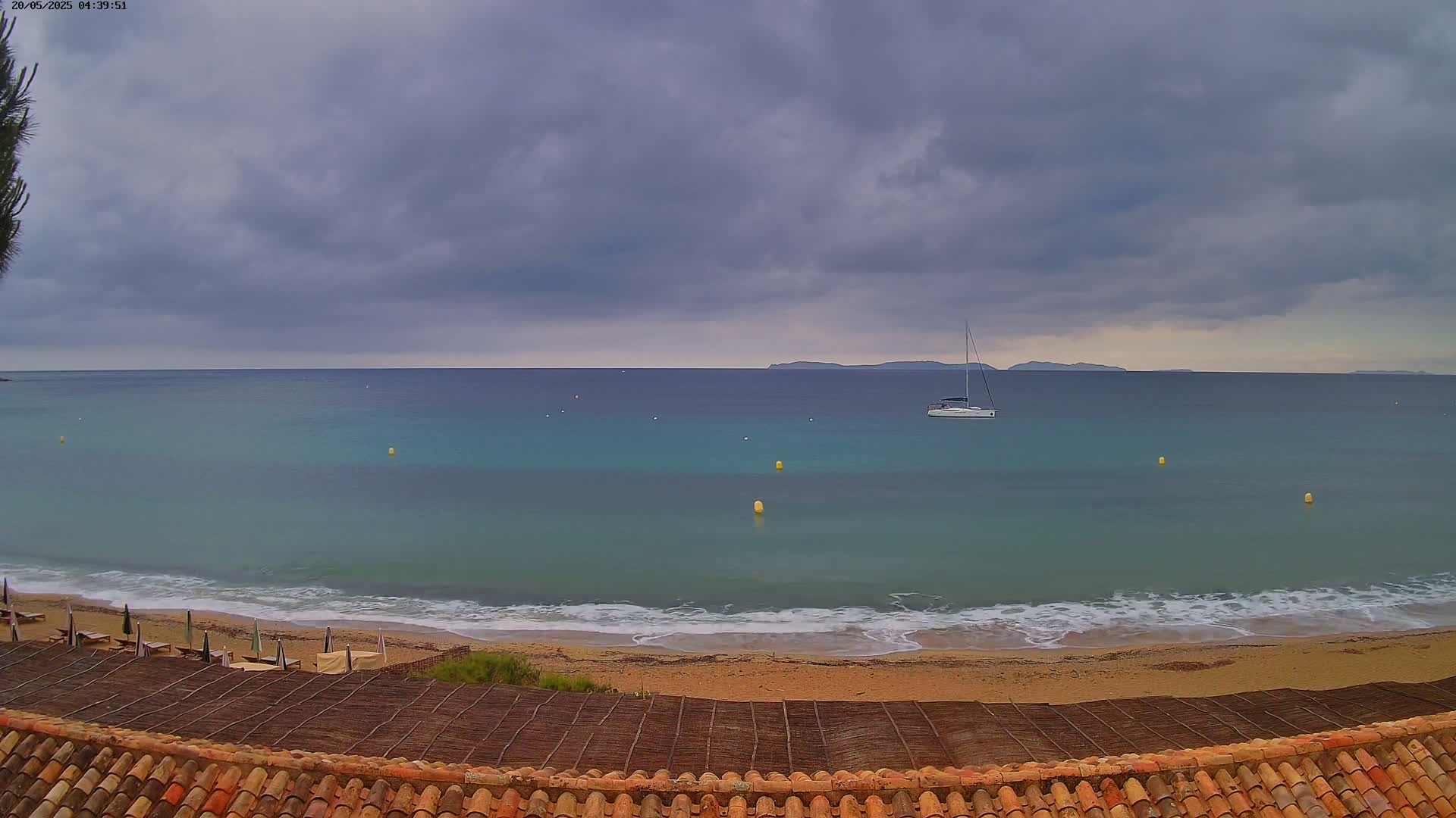 A sailboat sits on a calm, teal sea under a cloudy sky, viewed from above a sandy beach with beach umbrellas and a tiled roof.