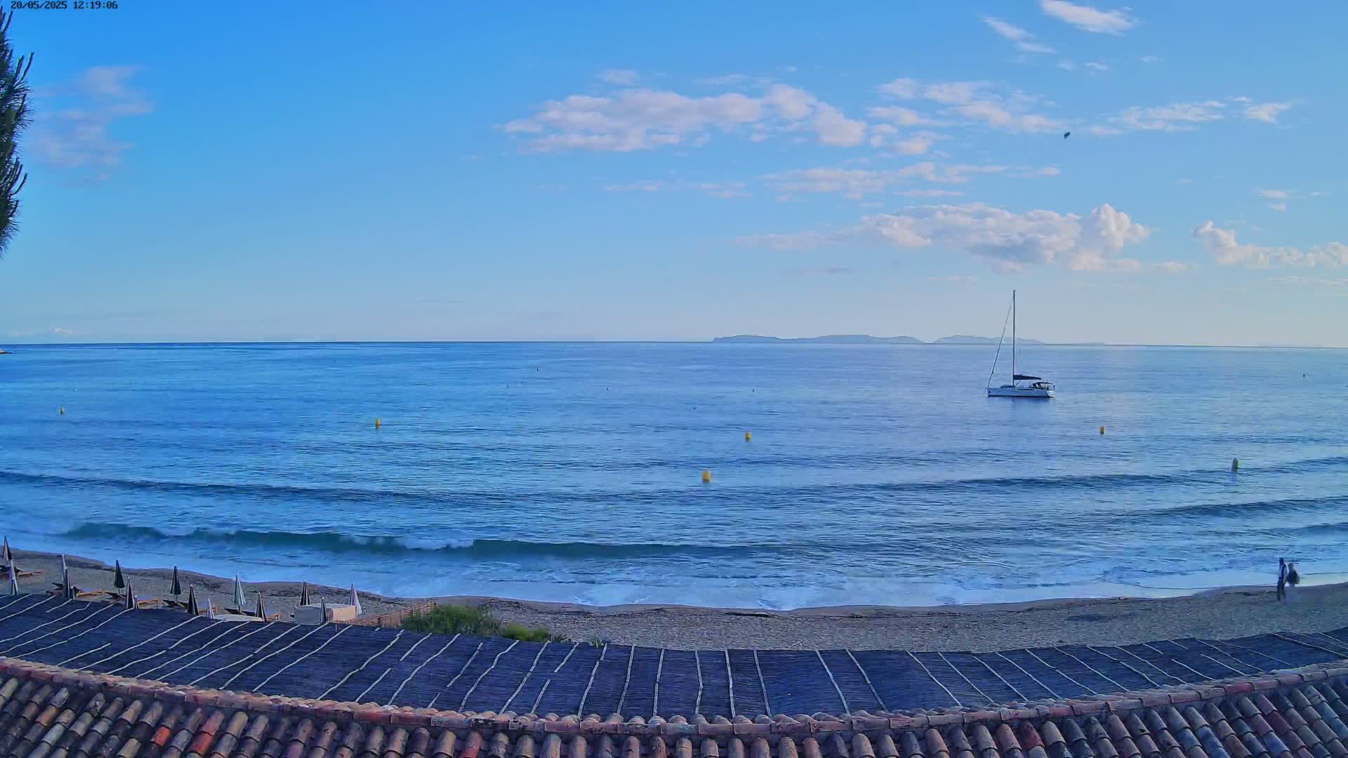 A sailboat sits on a calm, blue ocean under a mostly clear sky, with a sandy beach and tiled roof in the foreground.