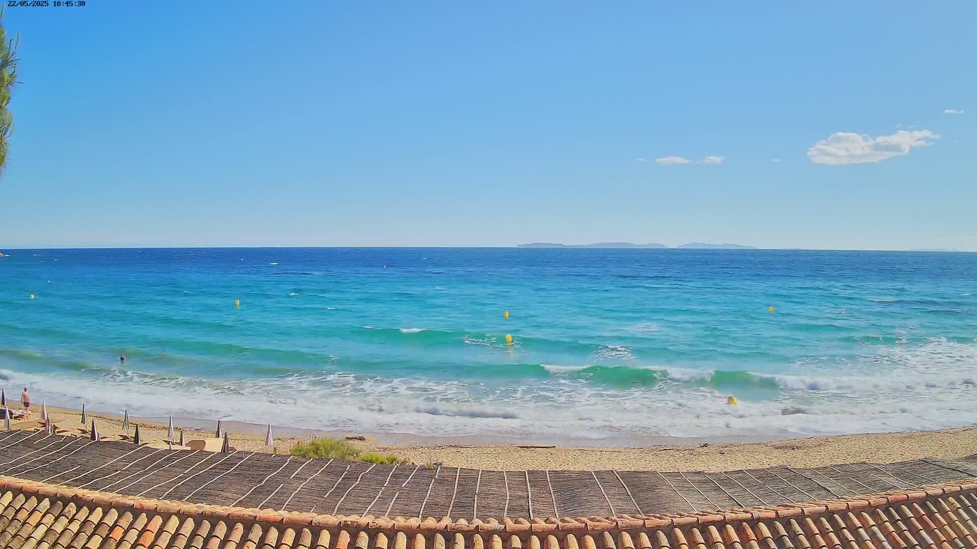 A sandy beach with a few people and umbrellas is visible from a rooftop with terracotta tiles, overlooking a turquoise ocean with small waves under a mostly clear blue sky.
