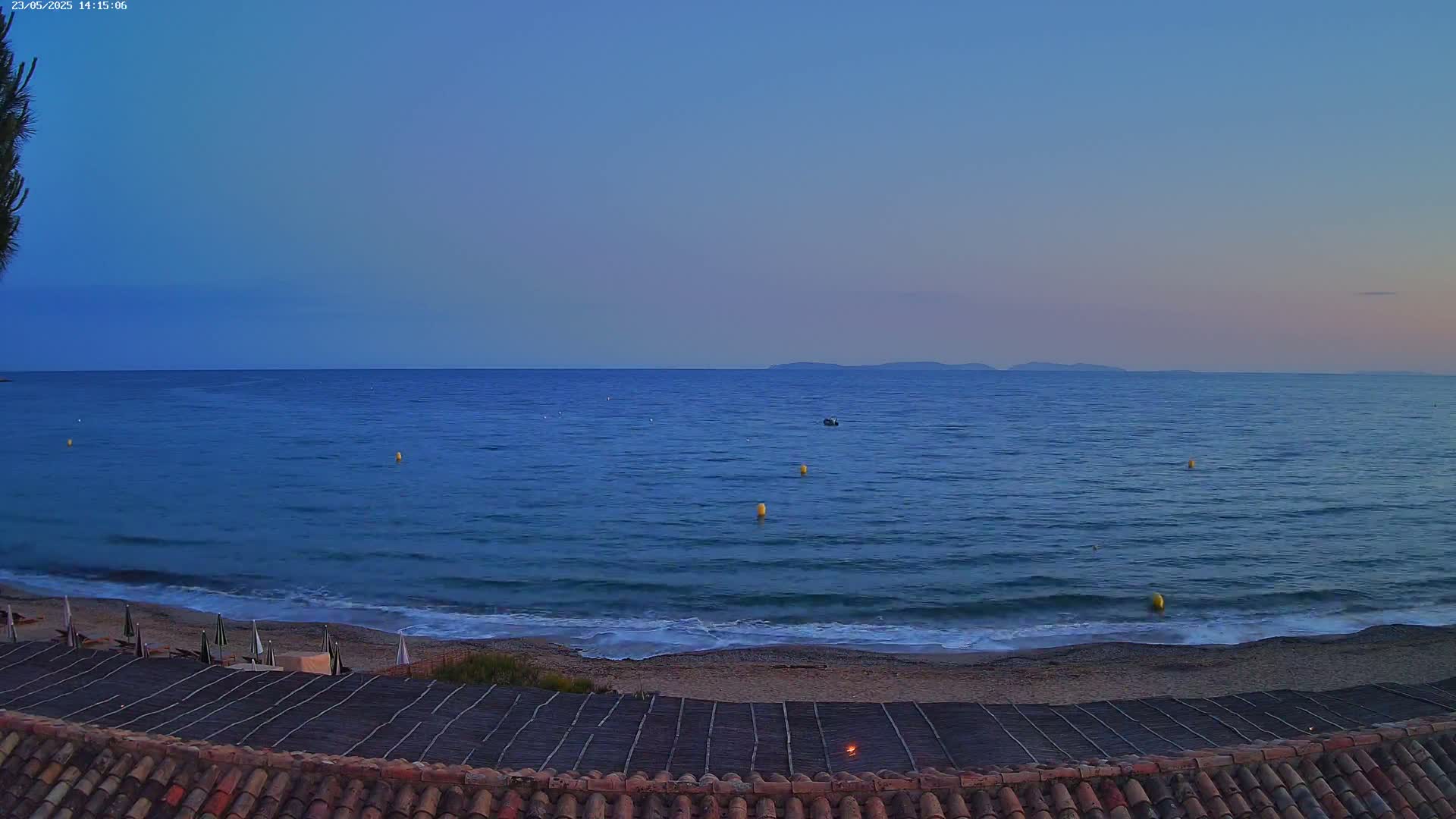 A calm, blue sea meets a sandy beach under a twilight sky, with a few yellow buoys marking the water.