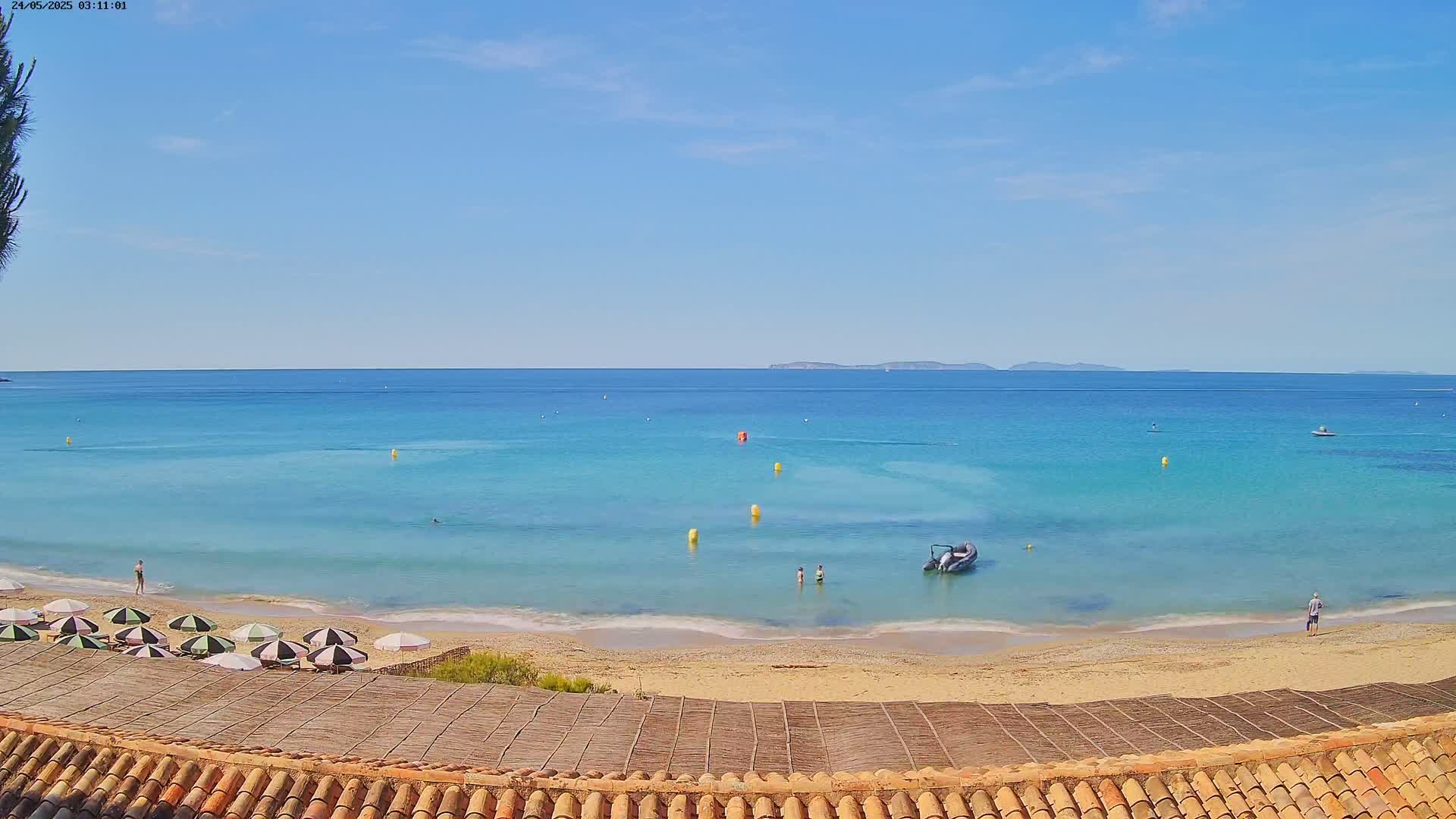 A sandy beach with a few people and striped umbrellas overlooks a calm, turquoise sea under a clear blue sky.