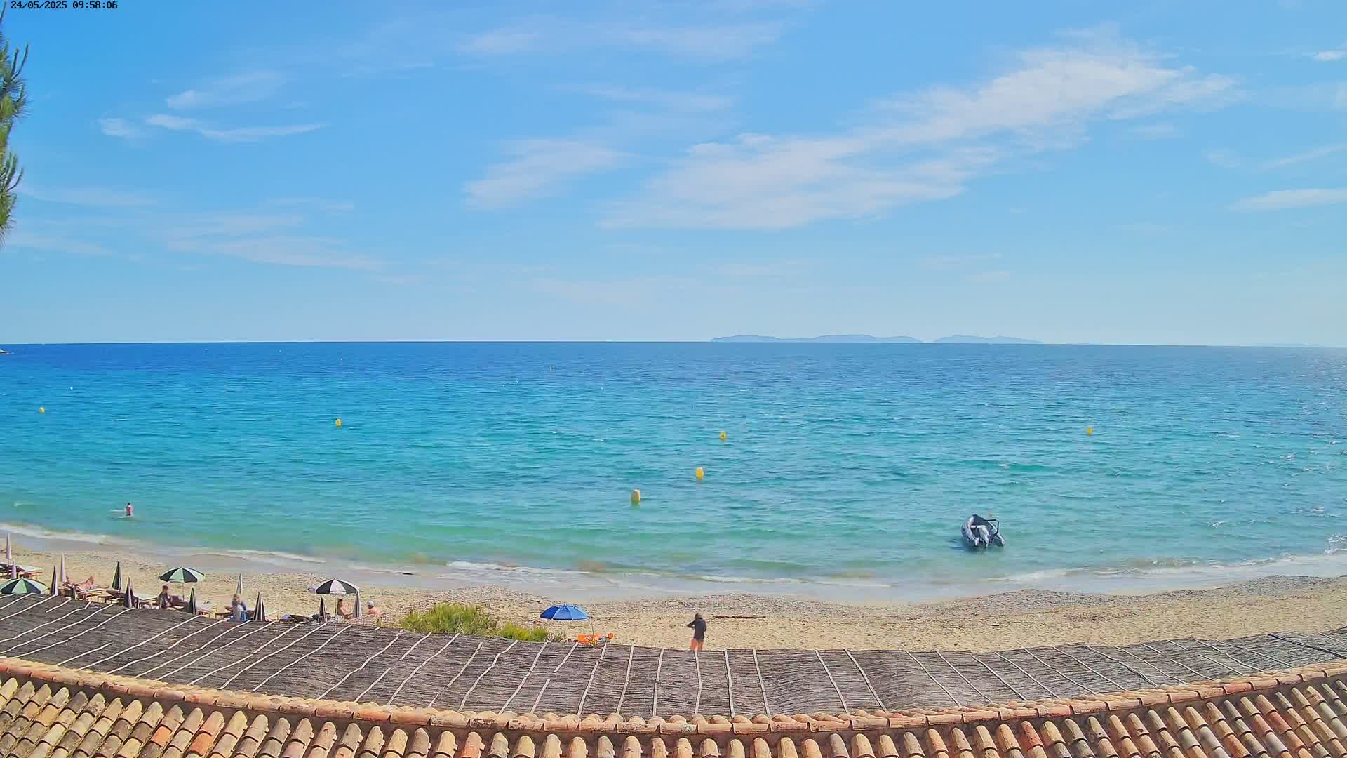A sandy beach with a few people and umbrellas is seen under a mostly sunny sky, overlooking a calm, turquoise ocean with a small boat and distant islands.