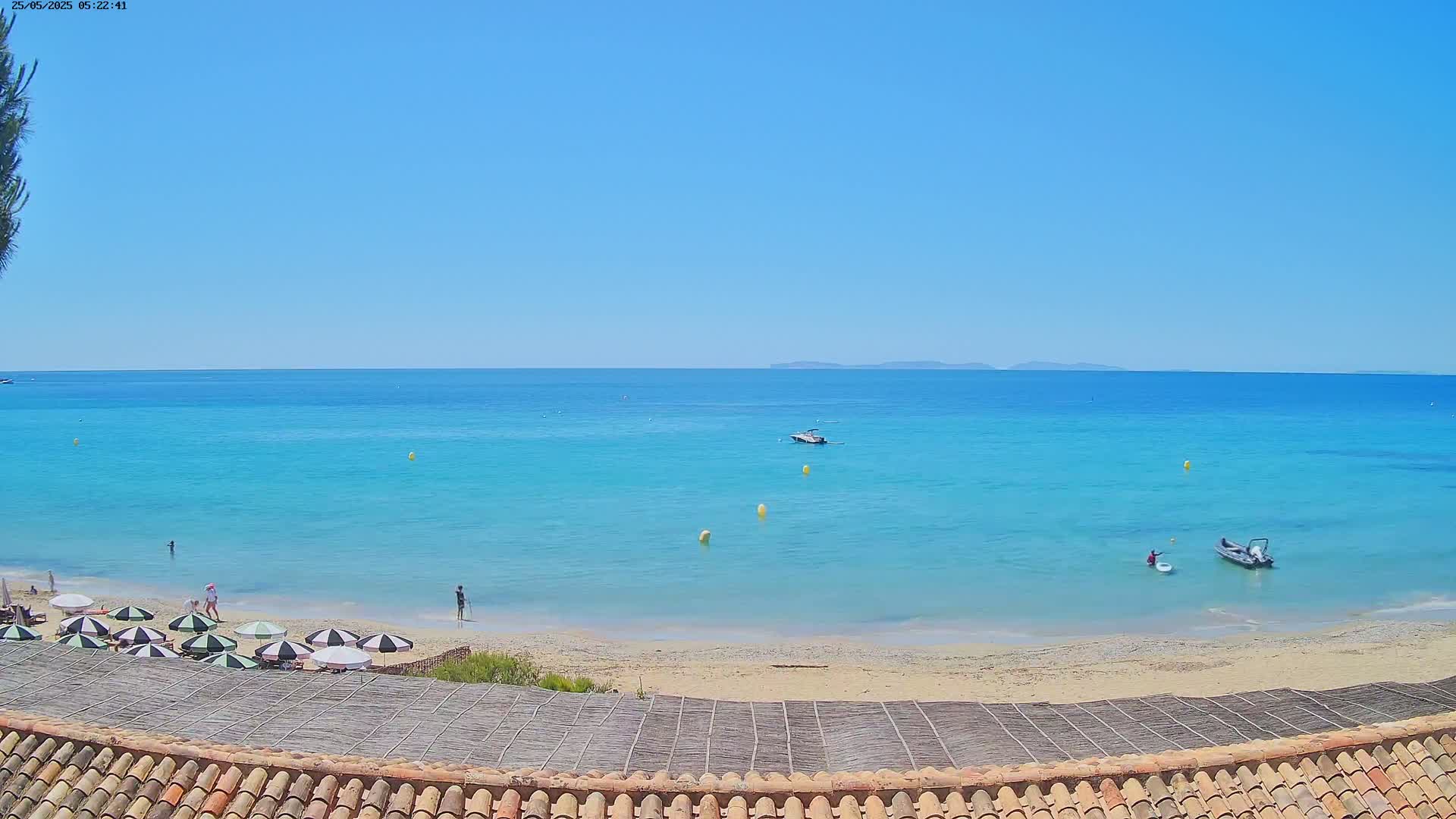 A sandy beach with turquoise water and several beach umbrellas under a clear blue sky is viewed from above a tiled roof.