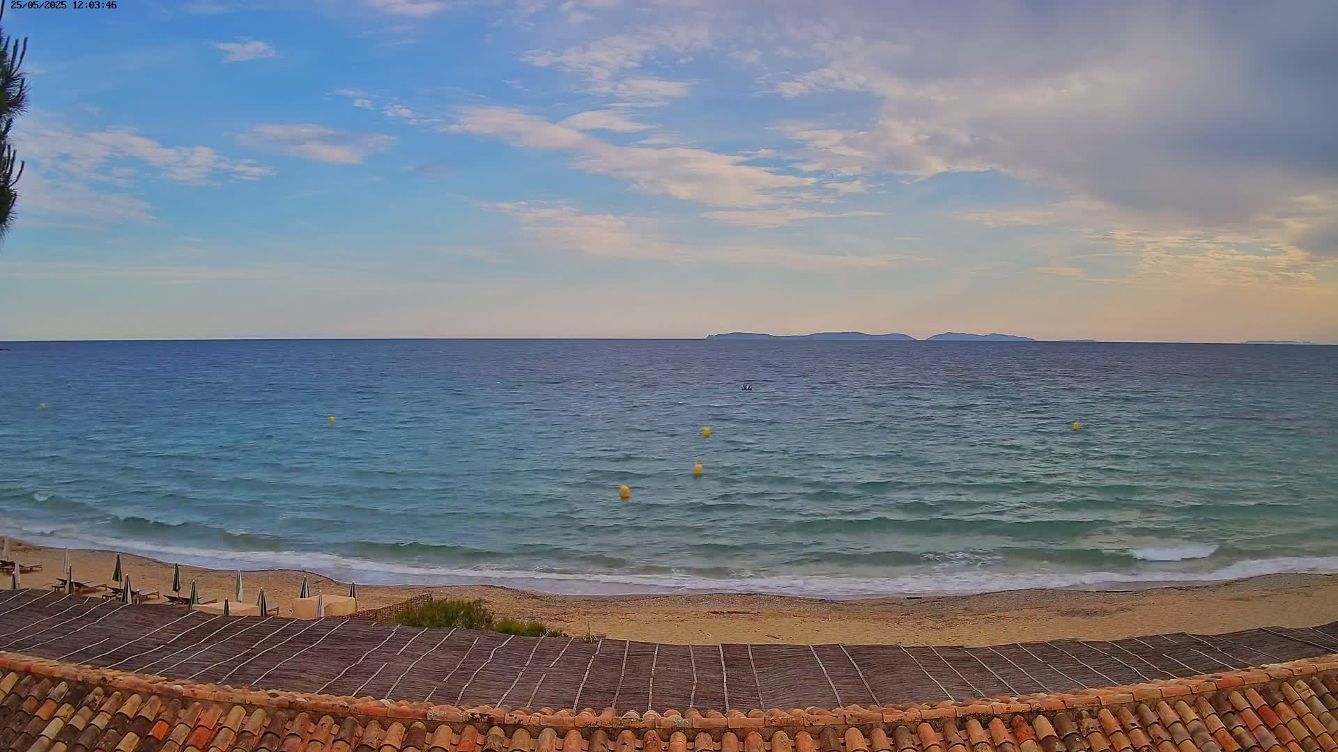 A partly cloudy sky overlooks a tranquil ocean beach with gently lapping waves, a sandy shore, and a thatched sun deck.
