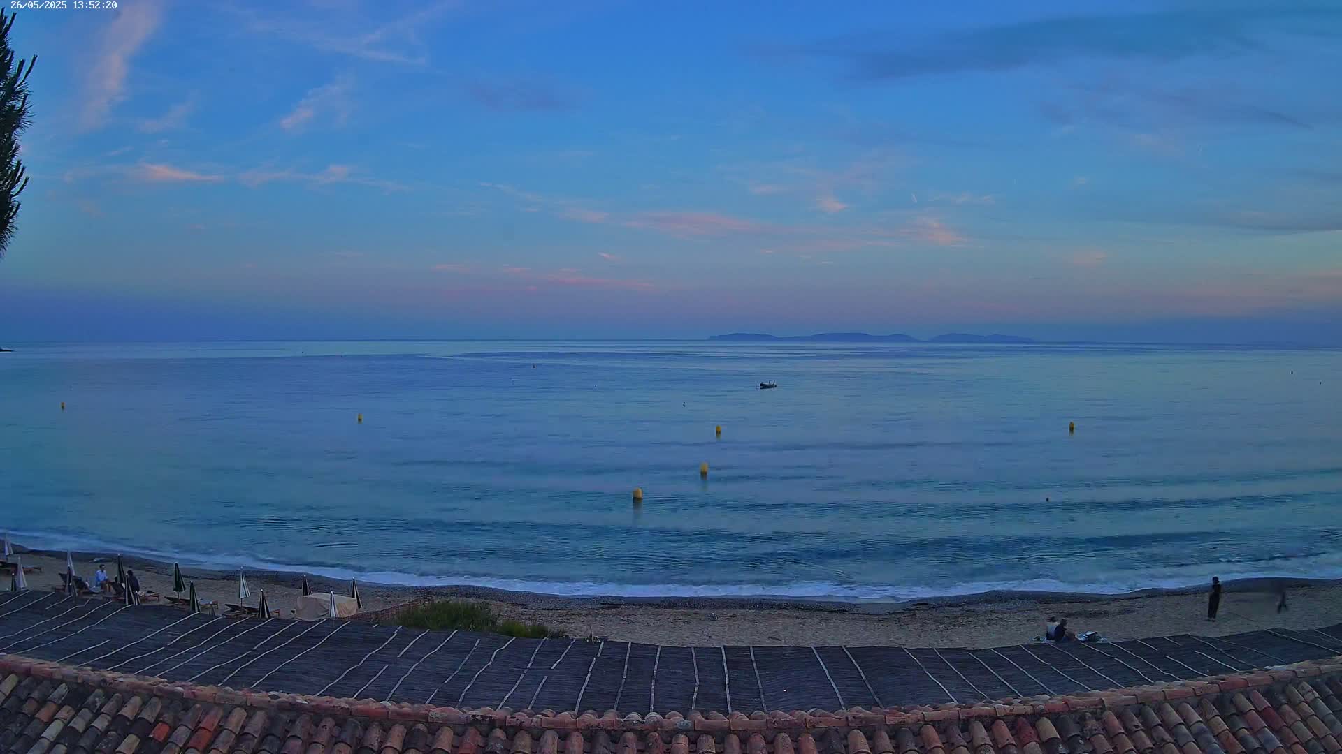 A calm, twilight ocean scene with a sandy beach, a few people, and some buoys in the water.