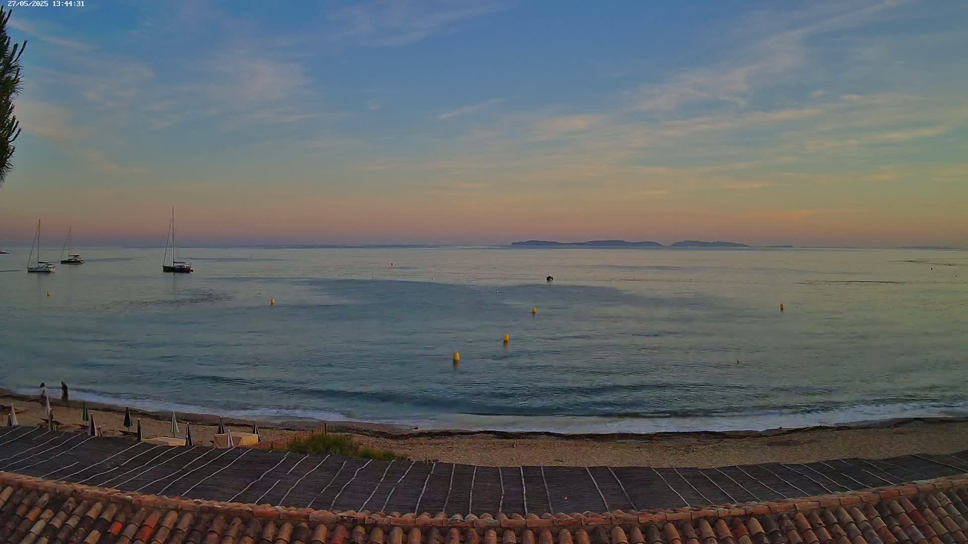 A calm sea at sunset, with a few sailboats anchored offshore and a sandy beach in the foreground.