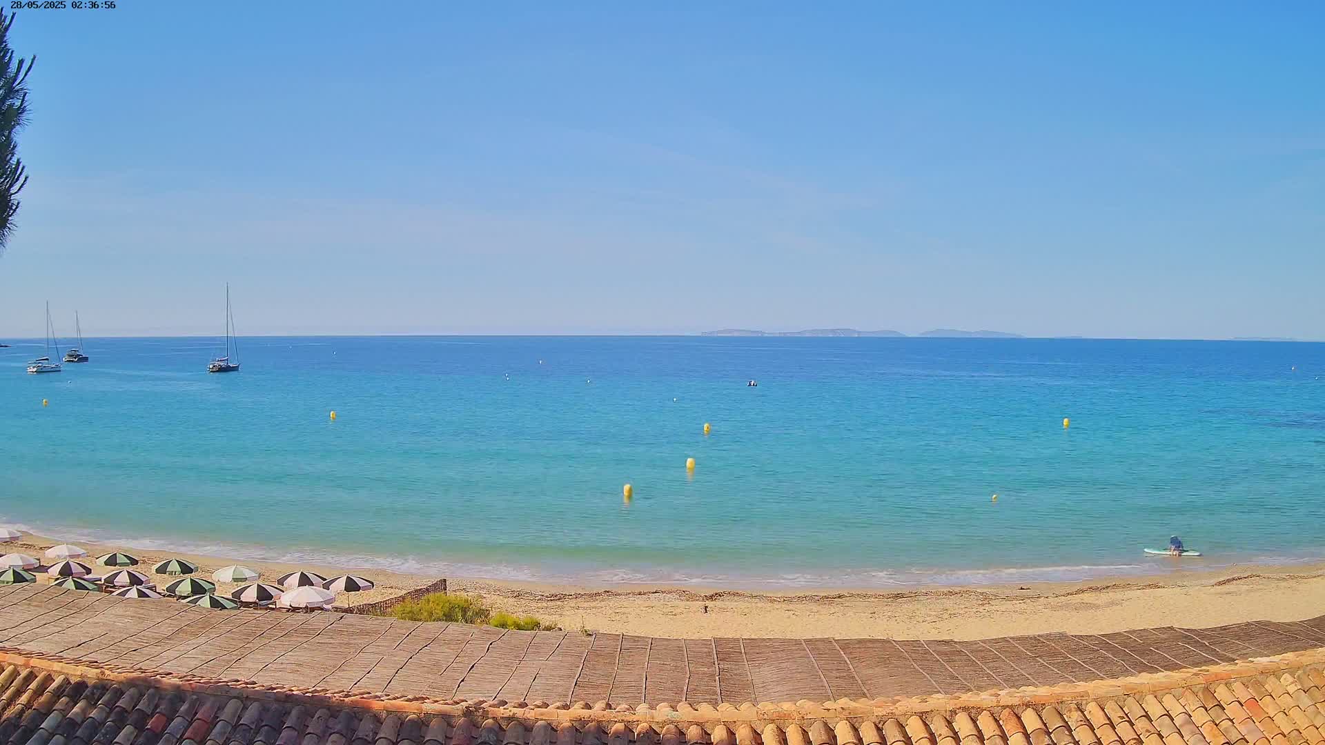 A sandy beach with several umbrellas is viewed from a tiled roof overlooking a calm, turquoise ocean with sailboats and a person on a paddleboard under a clear blue sky.