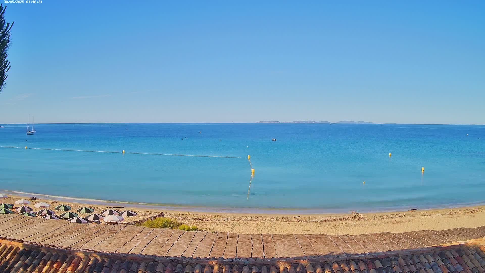 A sandy beach with striped umbrellas overlooks a calm, turquoise sea under a clear blue sky.