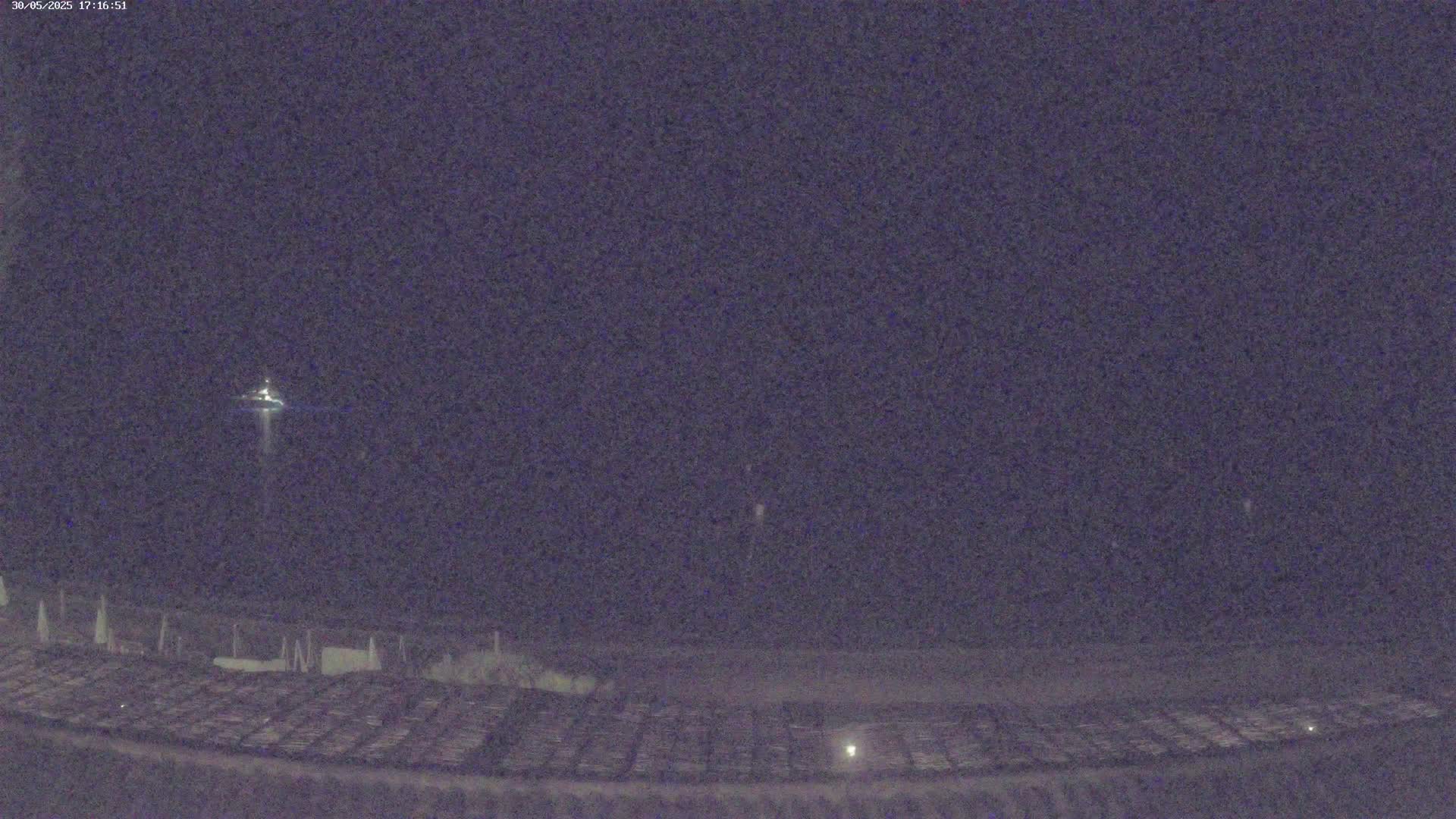 A nighttime long shot shows a small, distant light possibly from a boat on dark water, in front of a dark shoreline with a grid-like pattern.