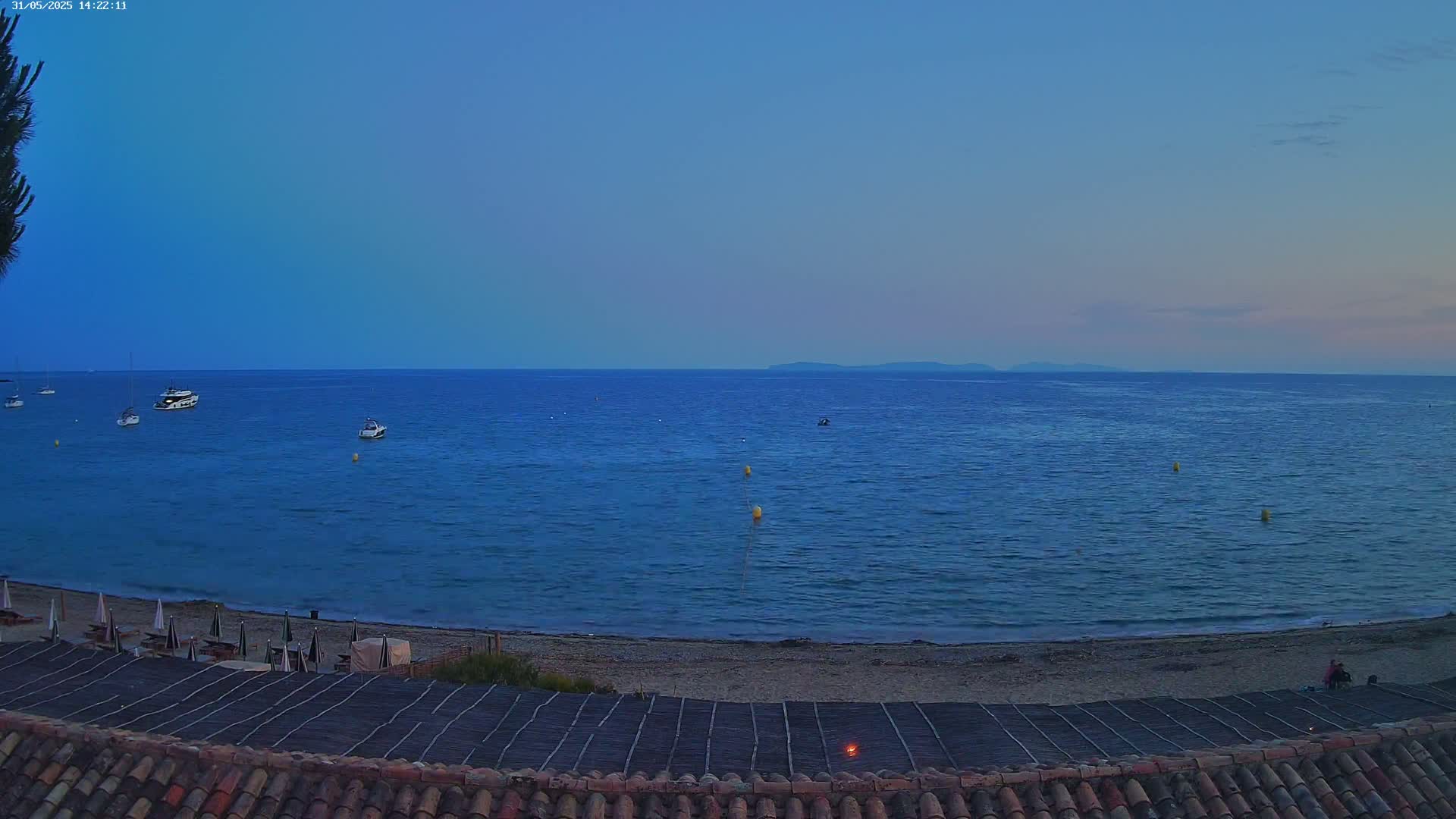 A tranquil beach scene at dusk shows a calm, dark-blue sea with several small boats, a sandy shore with beach umbrellas, and a tiled roof in the foreground.