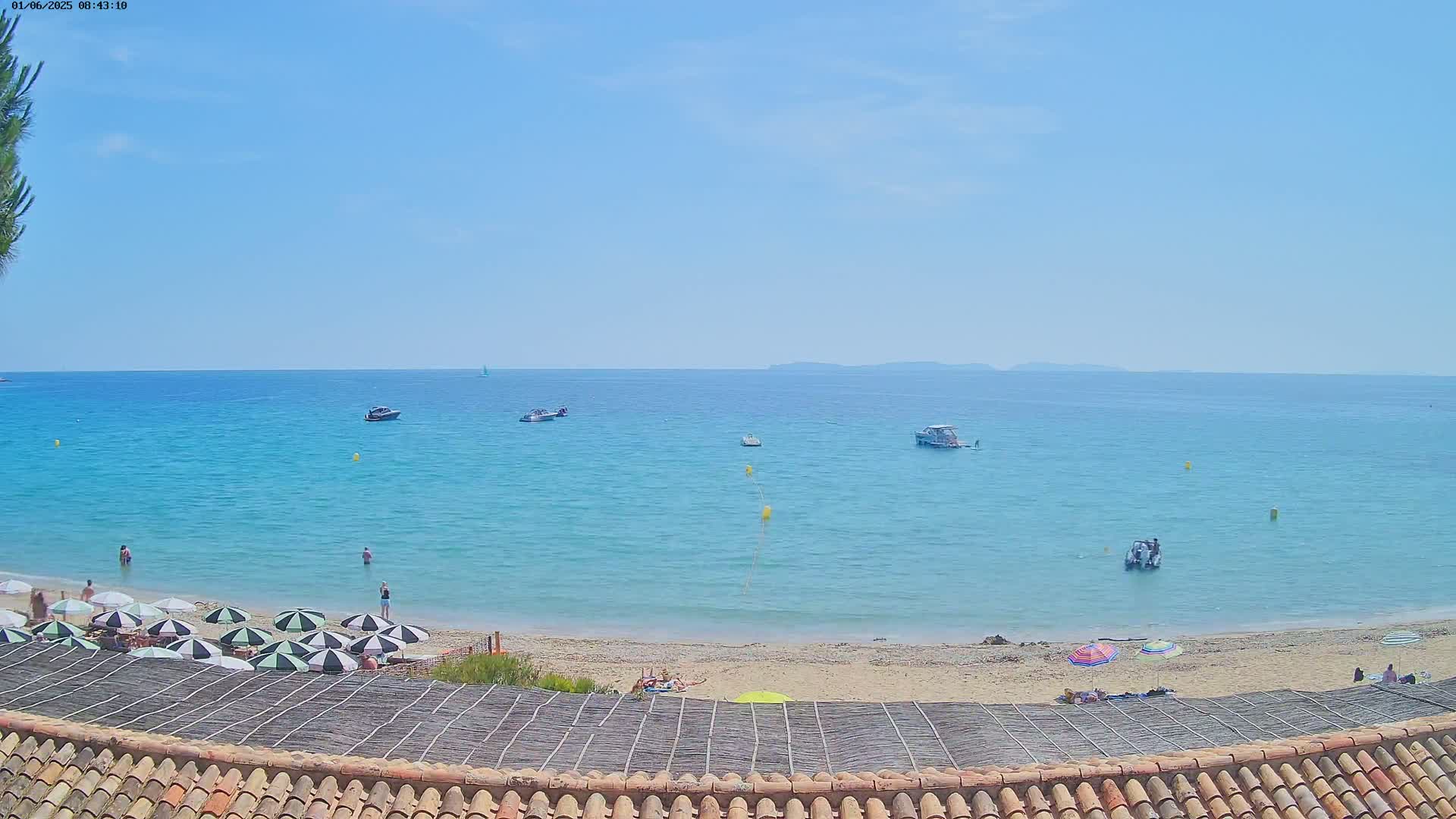 A sandy beach with several black and white striped umbrellas and a few people is visible, overlooking a calm, light-blue ocean with several small boats on a sunny day.