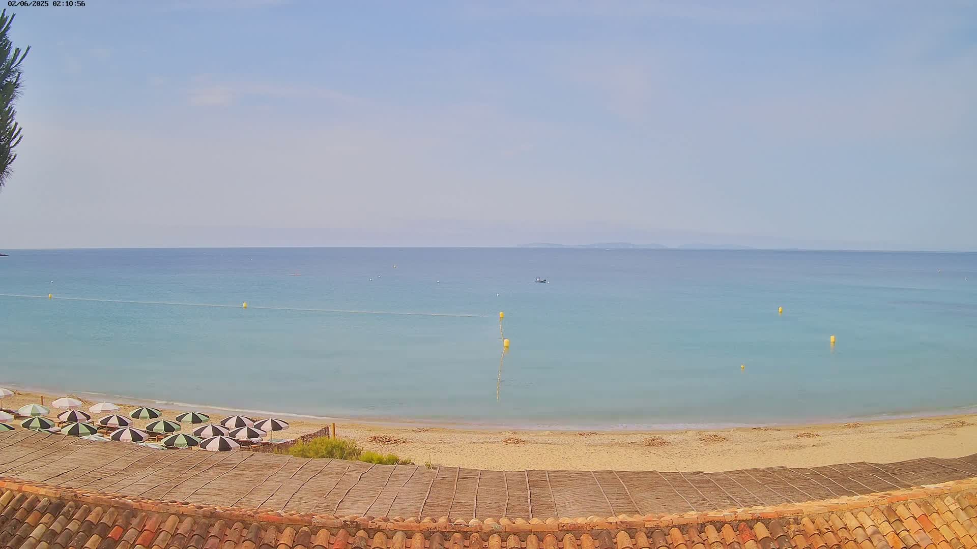 A calm, light-blue sea laps a sandy beach with several black and white striped umbrellas under a clear sky.