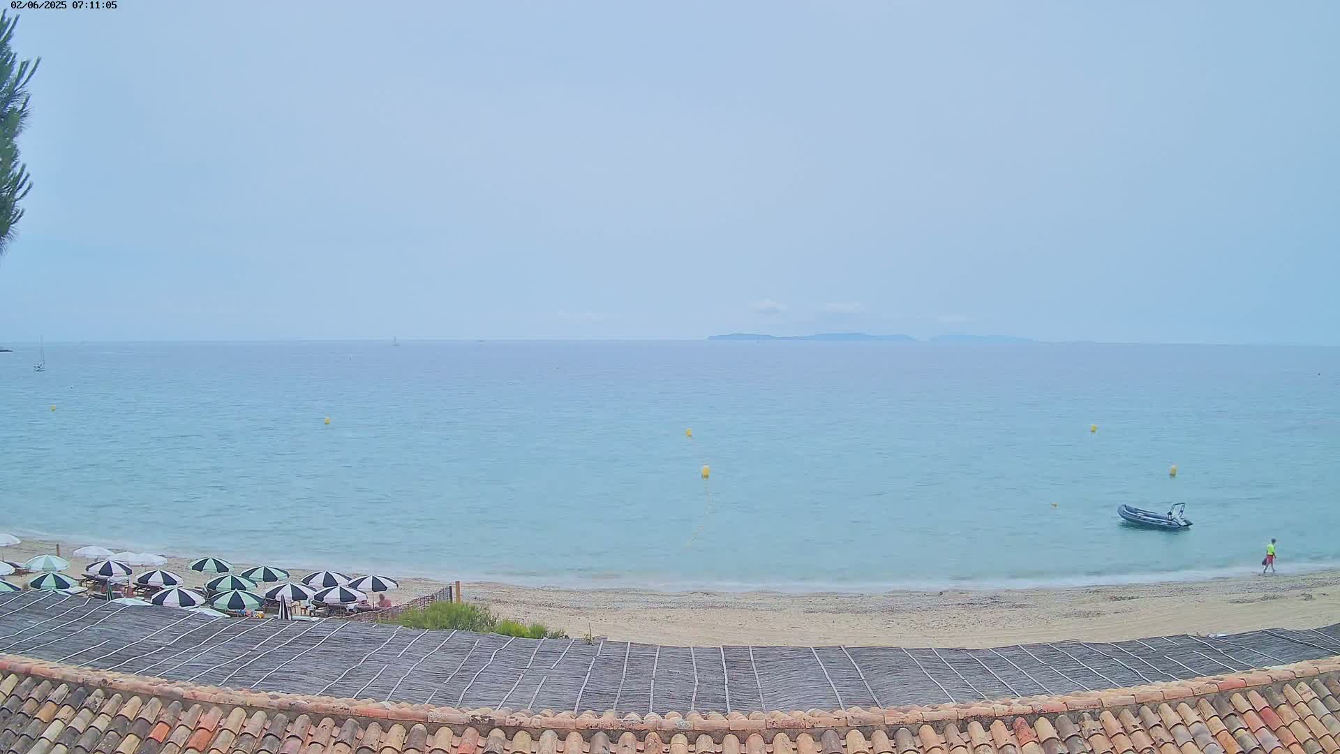 A calm, light-blue ocean stretches to a distant, low-lying island under a hazy sky, with a sandy beach featuring several closed umbrellas and a small inflatable boat visible in the foreground.