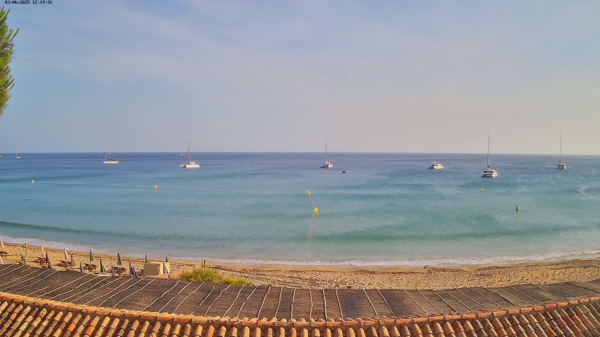 Several sailboats are anchored in calm, clear blue water off a sandy beach with beach umbrellas and a tiled roof in the foreground.