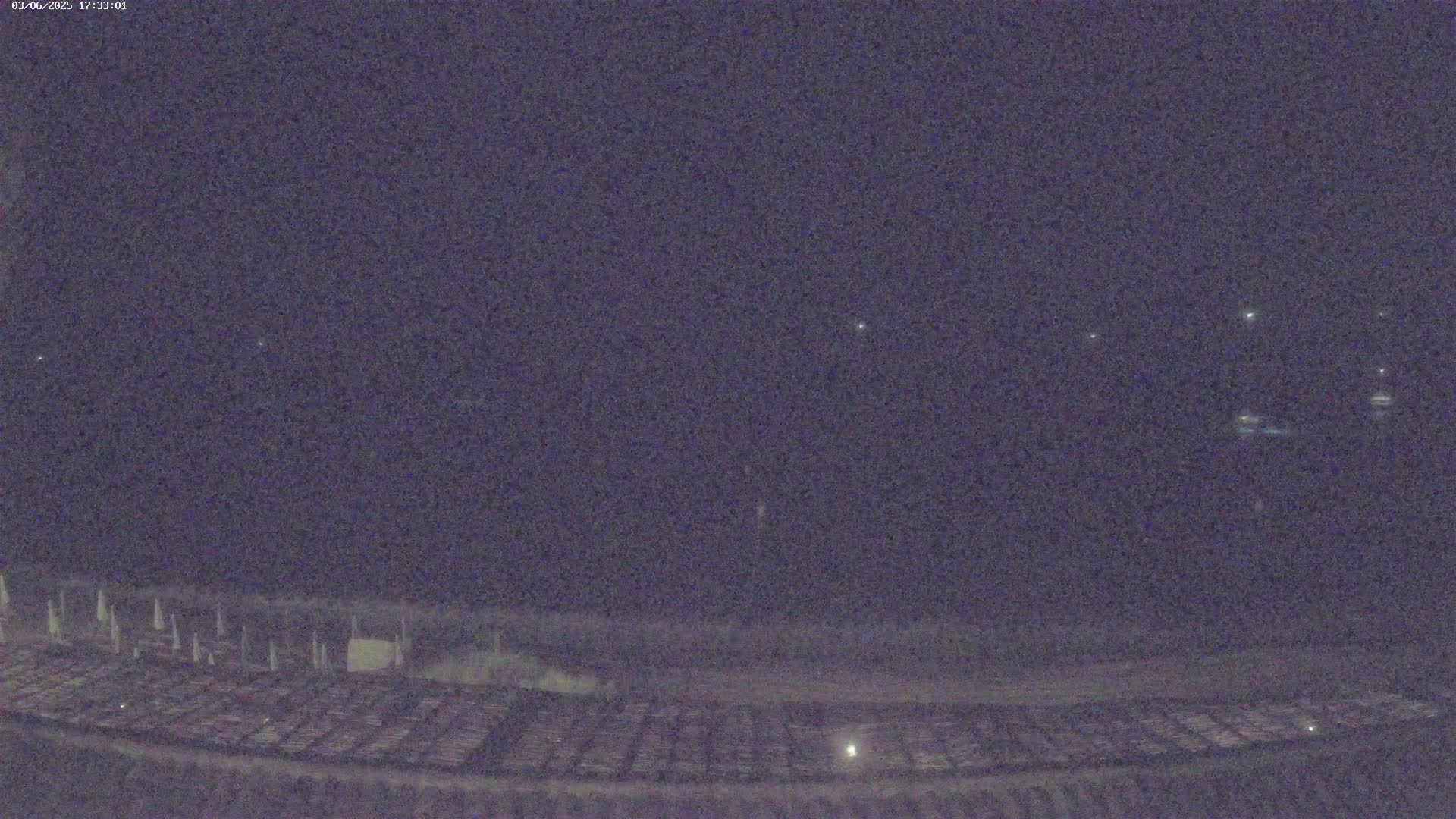 A nighttime, low-light view shows a dark, flat expanse of land with some faintly visible objects, possibly structures or vehicles, under a dark sky with a few distant lights.