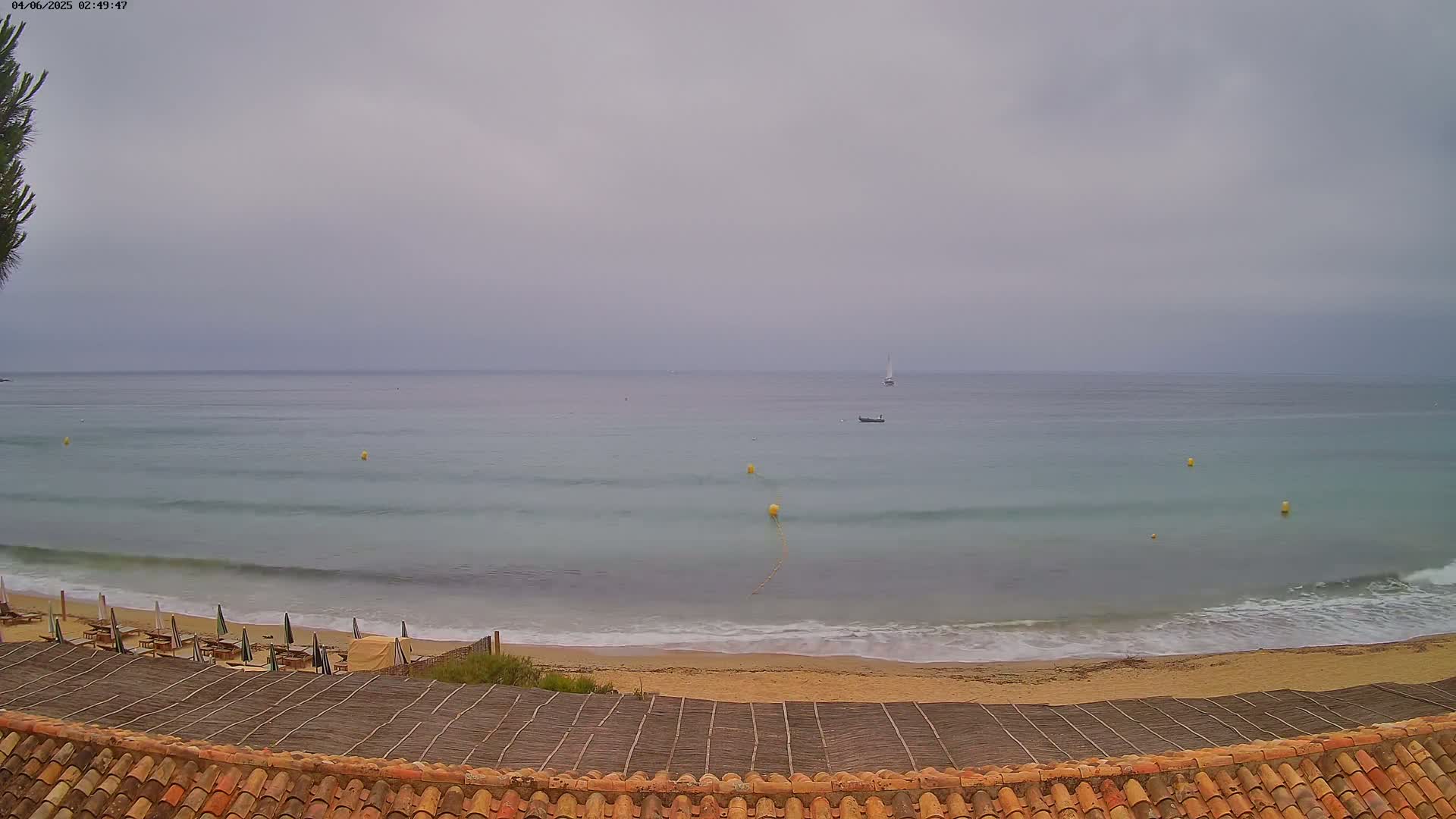 A beach with calm, light-grey water under an overcast sky, shows a few small boats offshore and sun umbrellas on the sand.