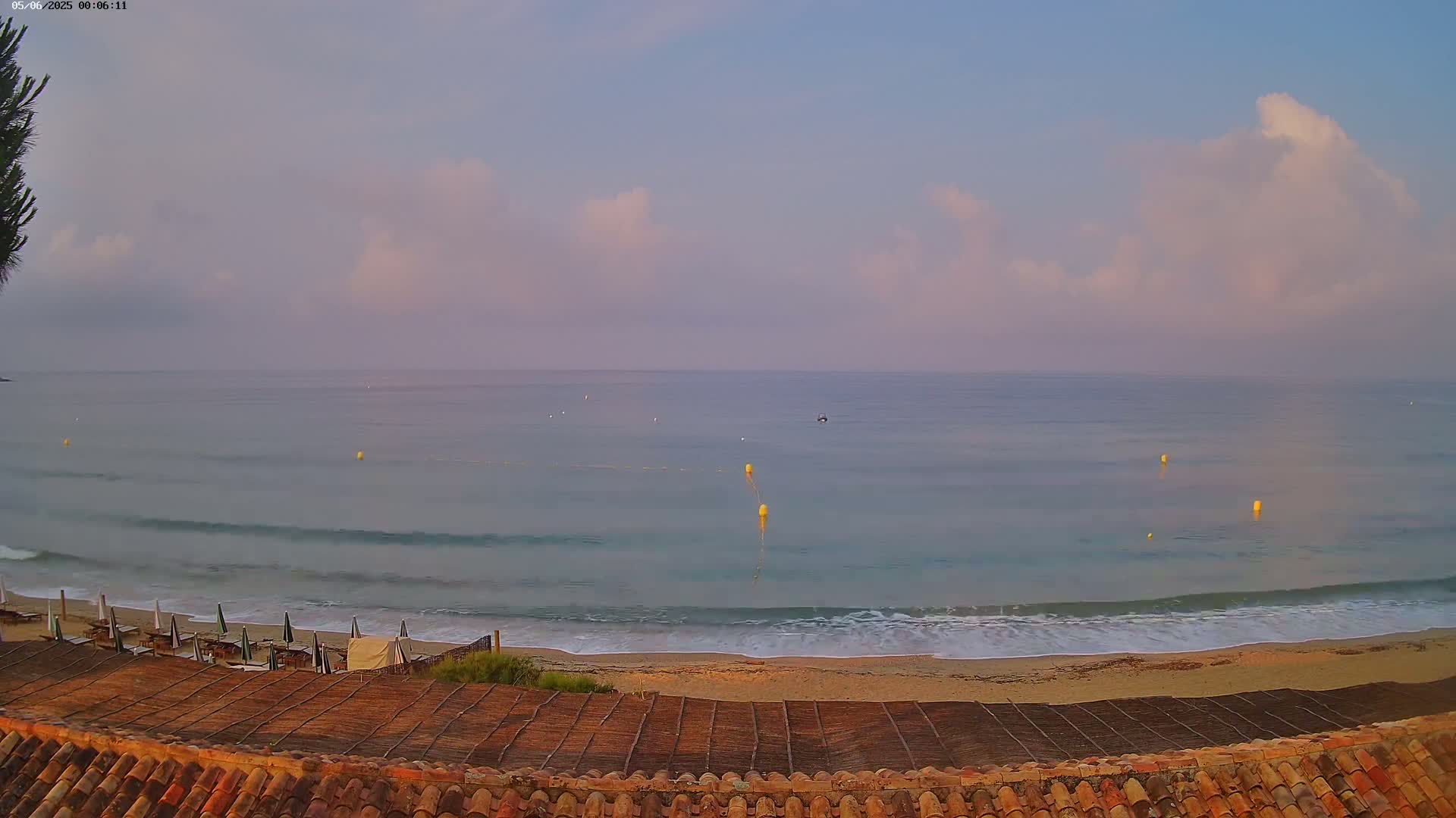 A calm ocean with small waves laps a sandy beach under a partly cloudy, pale sky at dawn.